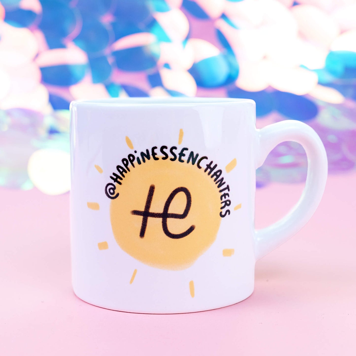 Close-up of a white mug with the Happiness Enchanters logo, showing a yellow sun with black text "@HappinessEnchanters" above it. The mug's handle is visible on the right side. The background consists of pastel hues, creating a dreamy atmosphere that enhances the bright, positive imagery on the mug.