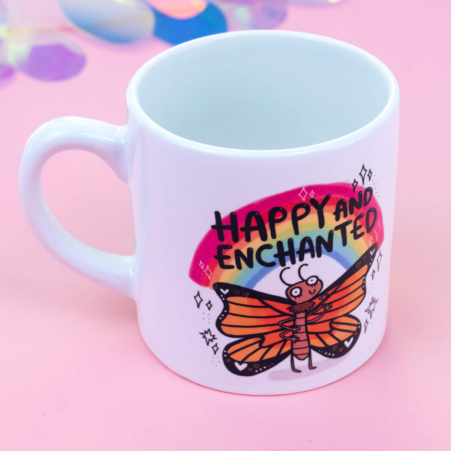 A white ceramic mug displaying a whimsical illustration of a colourful butterfly with a cheerful expression, beneath a vibrant rainbow. The text "Happy and Enchanted" appears above the butterfly in playful, bold letters. The pink background with bokeh effects complements the joyful theme of the design.