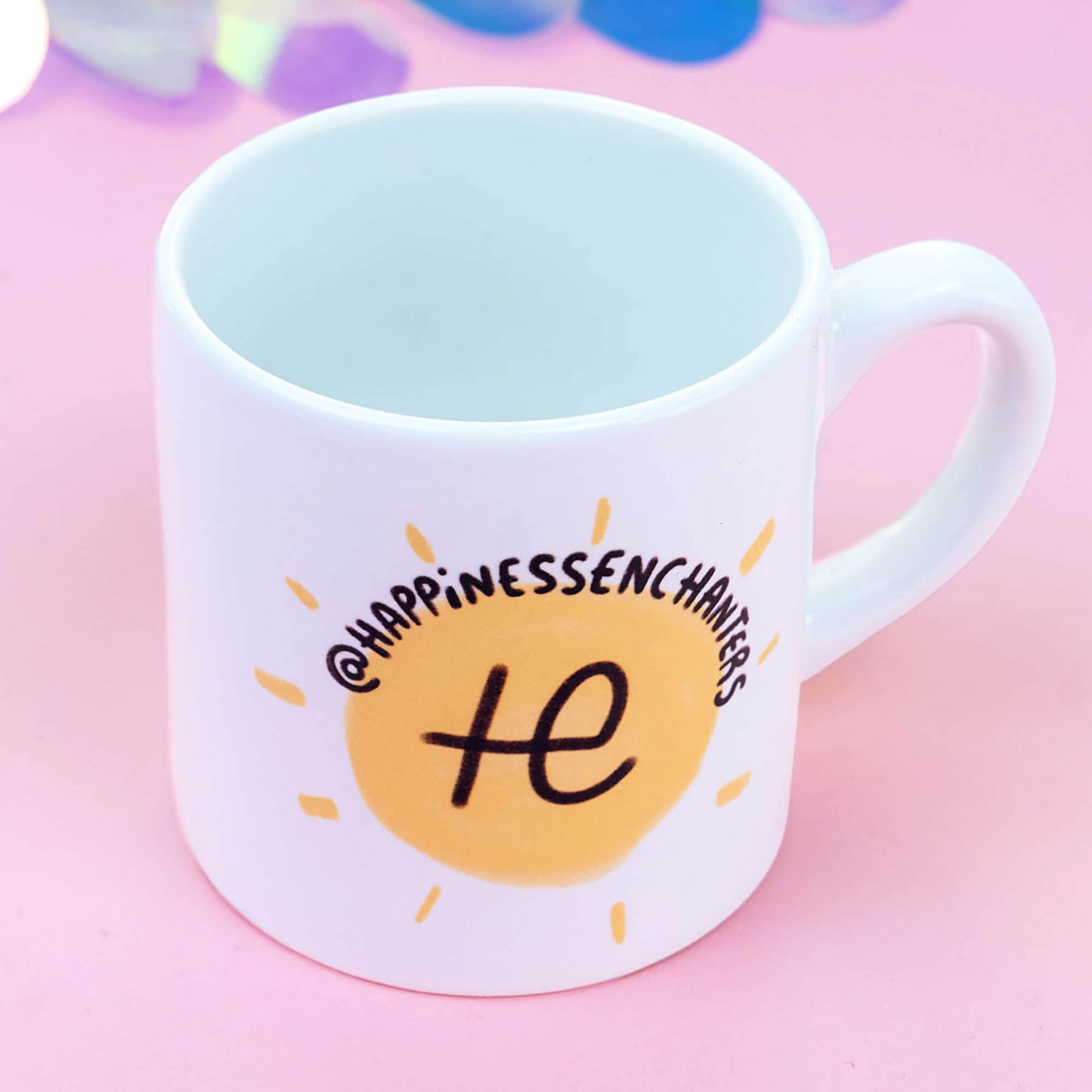 White ceramic mug featuring the Happiness Enchanters logo, a stylised sun with rays, and the text "@HappinessEnchanters" in black, curving around the top. The background is a soft pink with a subtle gradient effect, perfect for showcasing this uplifting design.