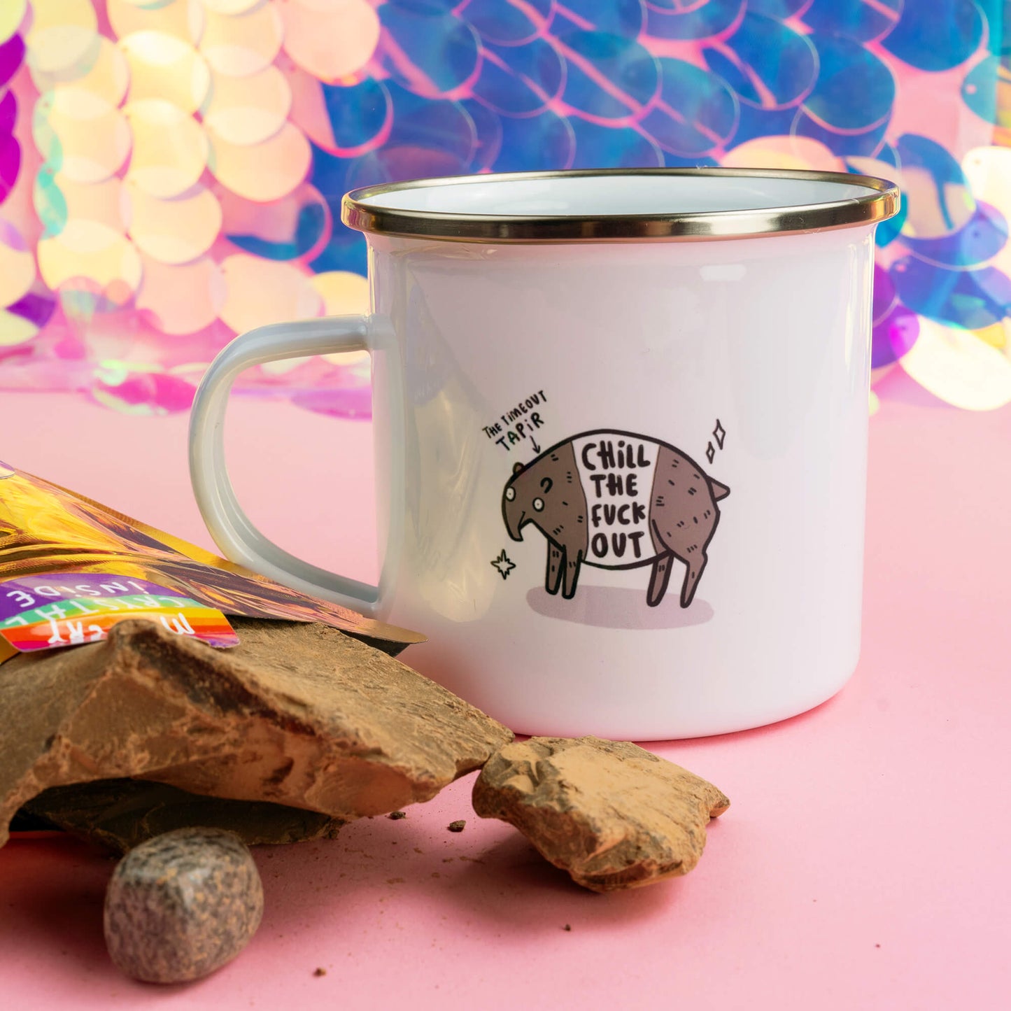 A white enamel mug with the phrase "Chill the F*** Out" beside a Happiness Enchanters gold pouch and cacao pieces on a pink background