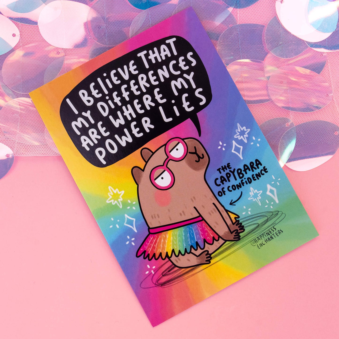 A close-up view of a postcard by 'Happiness Enchanters', displaying a charming cartoon capybara wearing pink glasses and a rainbow tutu, along with the phrase 'I believe that my differences are where my power lies'. The background is a bright rainbow gradient, capturing the essence of creativity and enchantment, with the caption 'The Capybara of Confidence'.