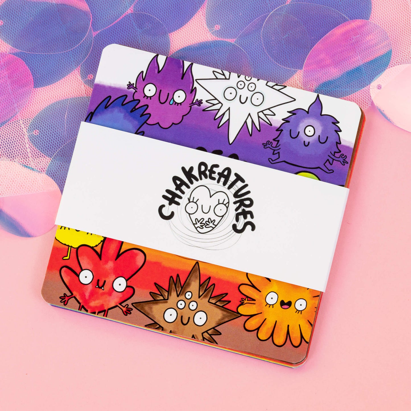 A bundle of Chakreatures cards by Happiness Enchanters, wrapped with a white band displaying the brand’s logo. The cards feature colourful, whimsical creatures symbolising different chakras, each with unique, playful expressions. The top cards display vibrant red, purple, and yellow designs on a mystical pink backdrop. Ideal for meditation, self-reflection, and connecting with spiritual energy through art.