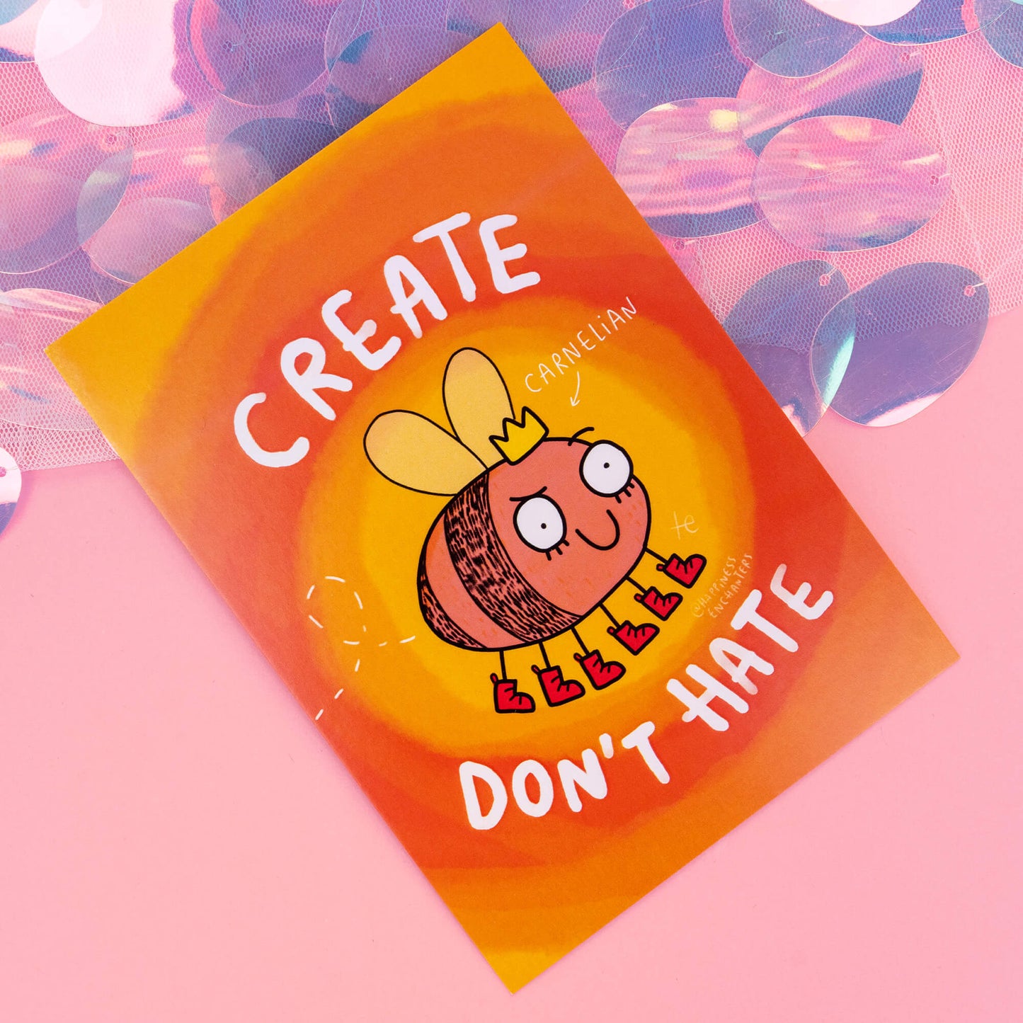 A close-up view of a Carnelian-themed postcard by 'Happiness Enchanters', displaying a charming cartoon Carnelian crystal bee with a crown and boots, along with the phrase 'Create, don't hate'. The background is a bright blend of orange and yellow with swirling patterns, capturing the essence of creativity and enchantment.