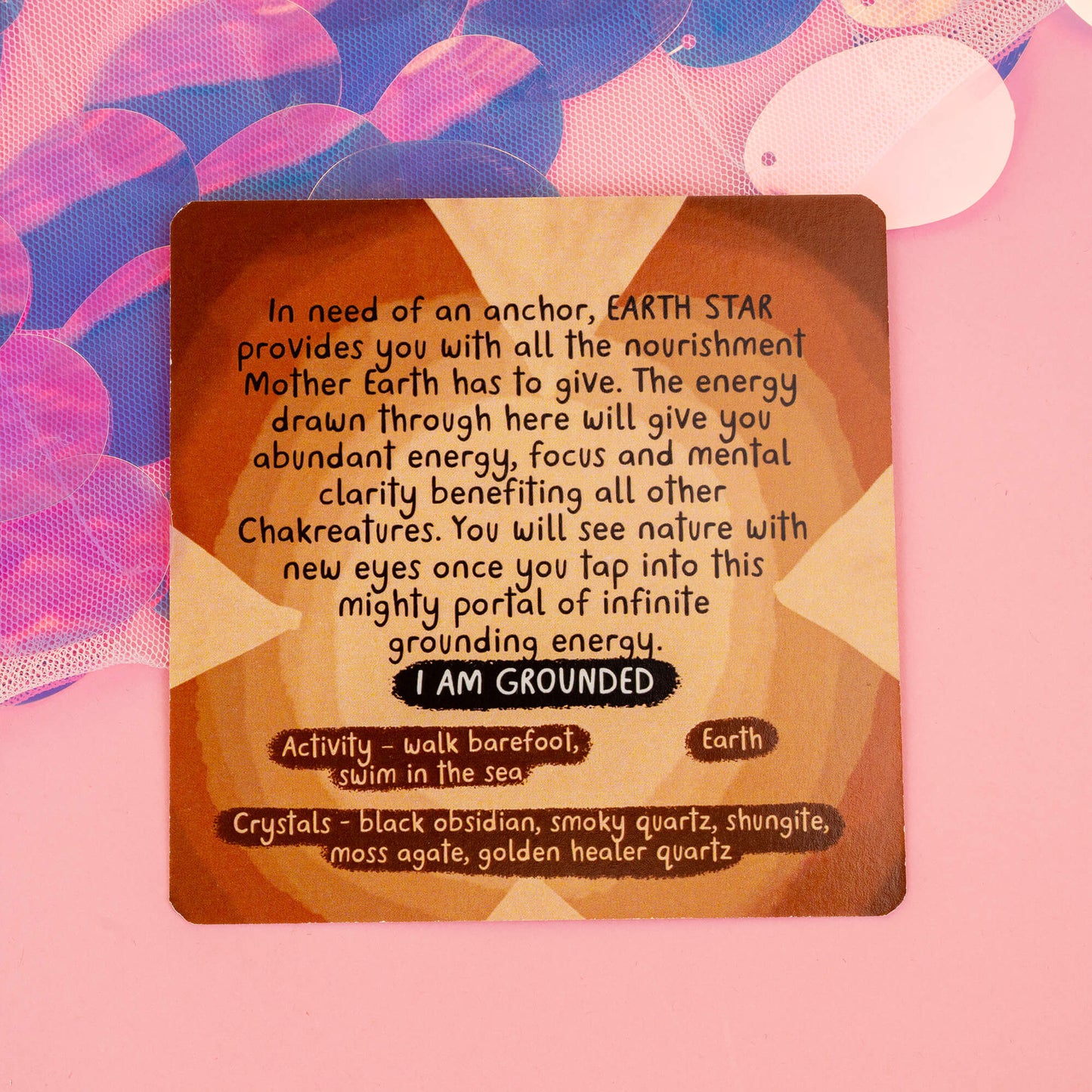 Earth Star chakra card from the Chakreatures deck, promoting grounding and connection with Mother Earth. The card suggests activities like walking barefoot and swimming, with grounding crystals like black obsidian and smoky quartz. Set against a pink background