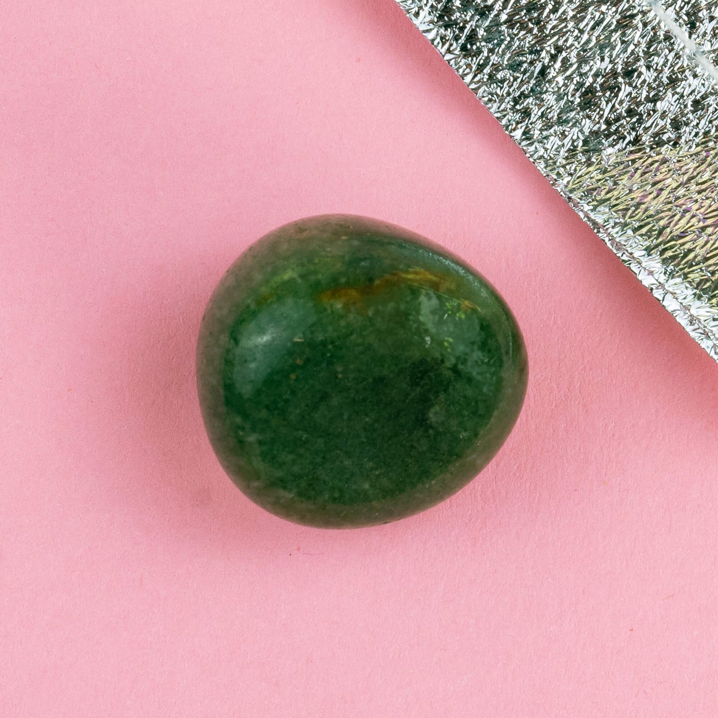 Happiness Enchanters green aventurine crystal on a pink backdrop with metallic silver bag laid next to it