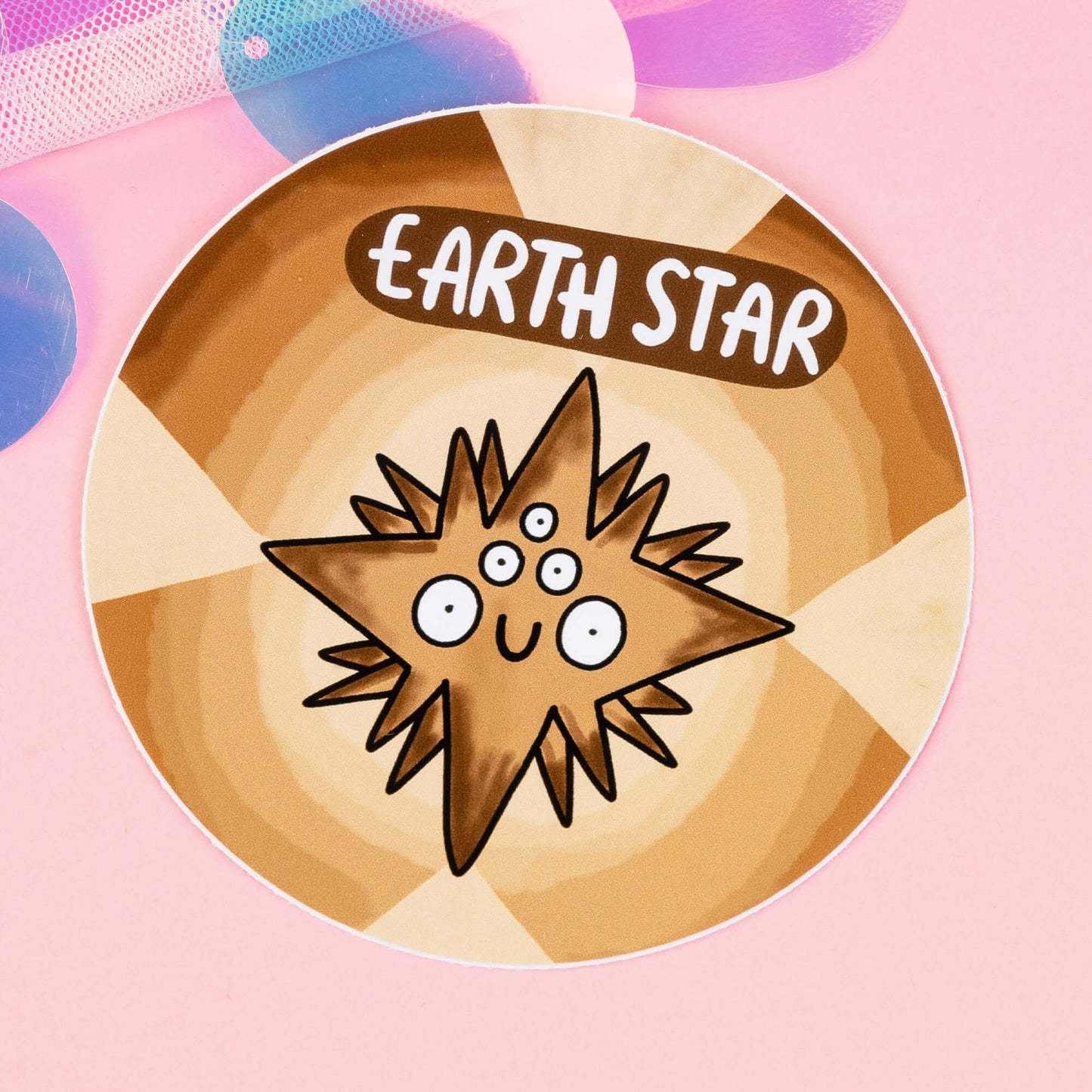 Earth Star Chakreatures sticker by Happiness Enchanters. A circular brown and beige sticker with a brown diamond shaped creature illustrated by Katie Abey with multiple eyes and smiling mouth. 'Earth Star' is written above the character. The sticker is on a pink backdrop.
