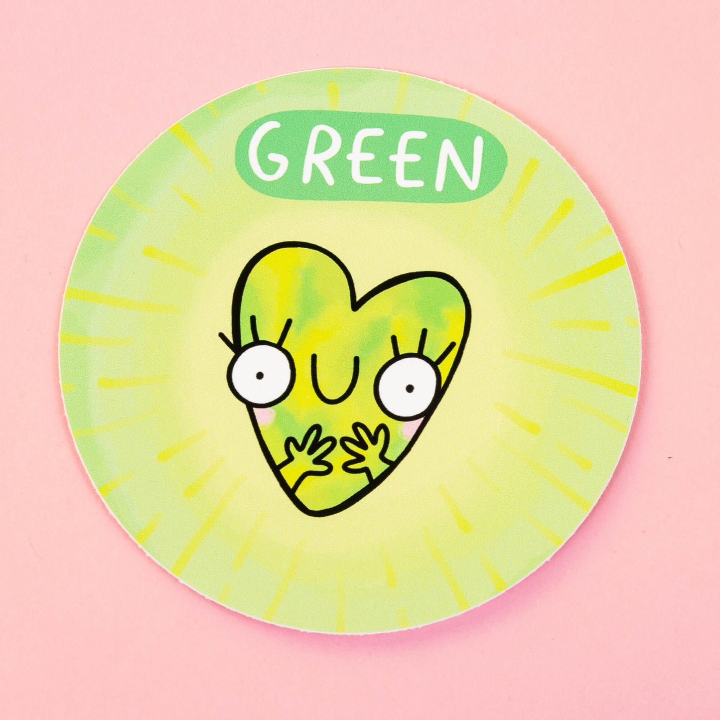 Green Chakreatures sticker by Happiness Enchanters. A circular green sticker with wide eyed, smiley heart shaped cartoon creature with blushing cheeks illustrated by Katie Abey. 'Green' is written above the character. The sticker is on a pink backdrop.