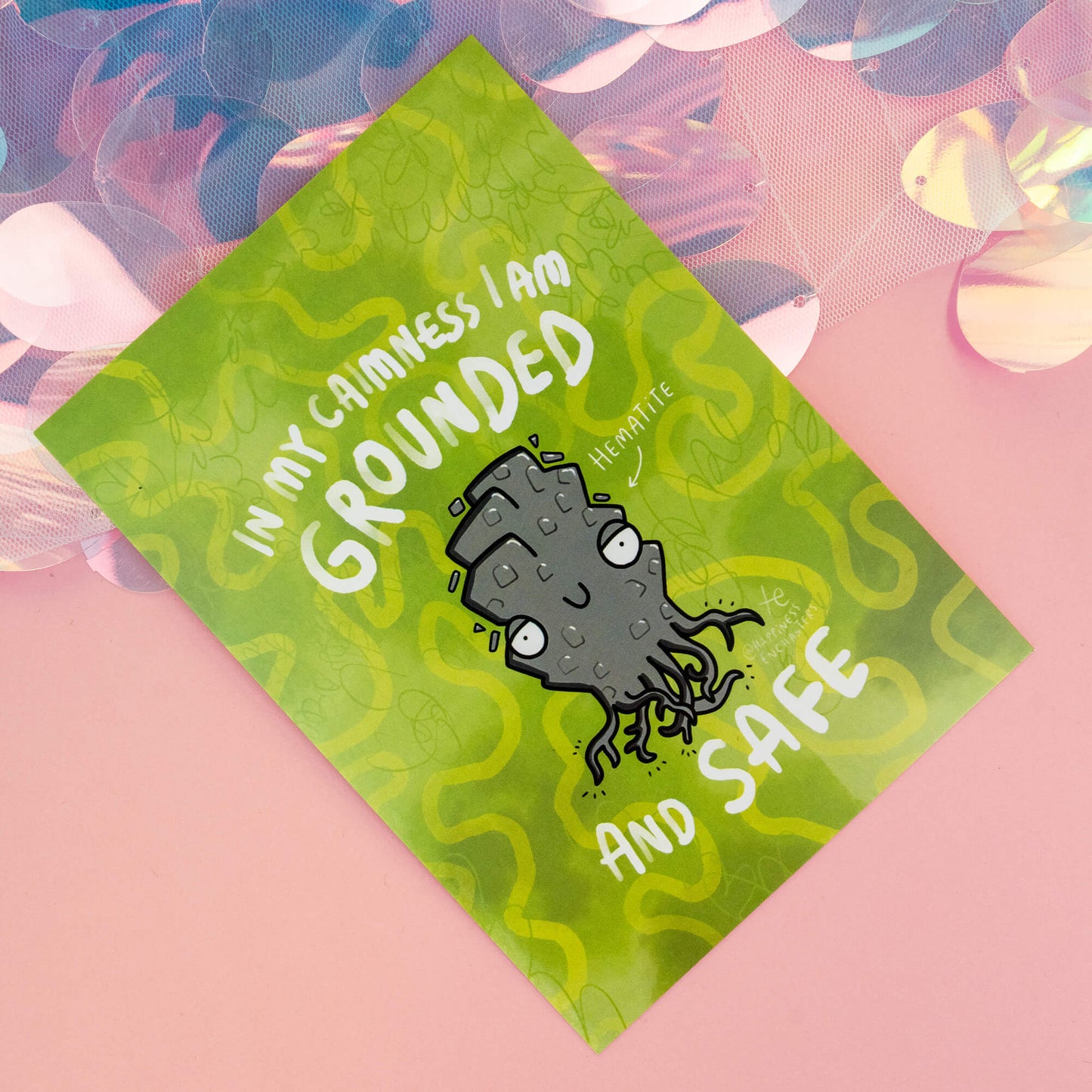 A whimsical Hematite-themed postcard from 'Happiness Enchanters', featuring a cute, cartoon Hematite crystal character with the text 'In my calmness I am grounded and safe' on a green, wavy-patterned background. The postcard exudes a magical and playful vibe.