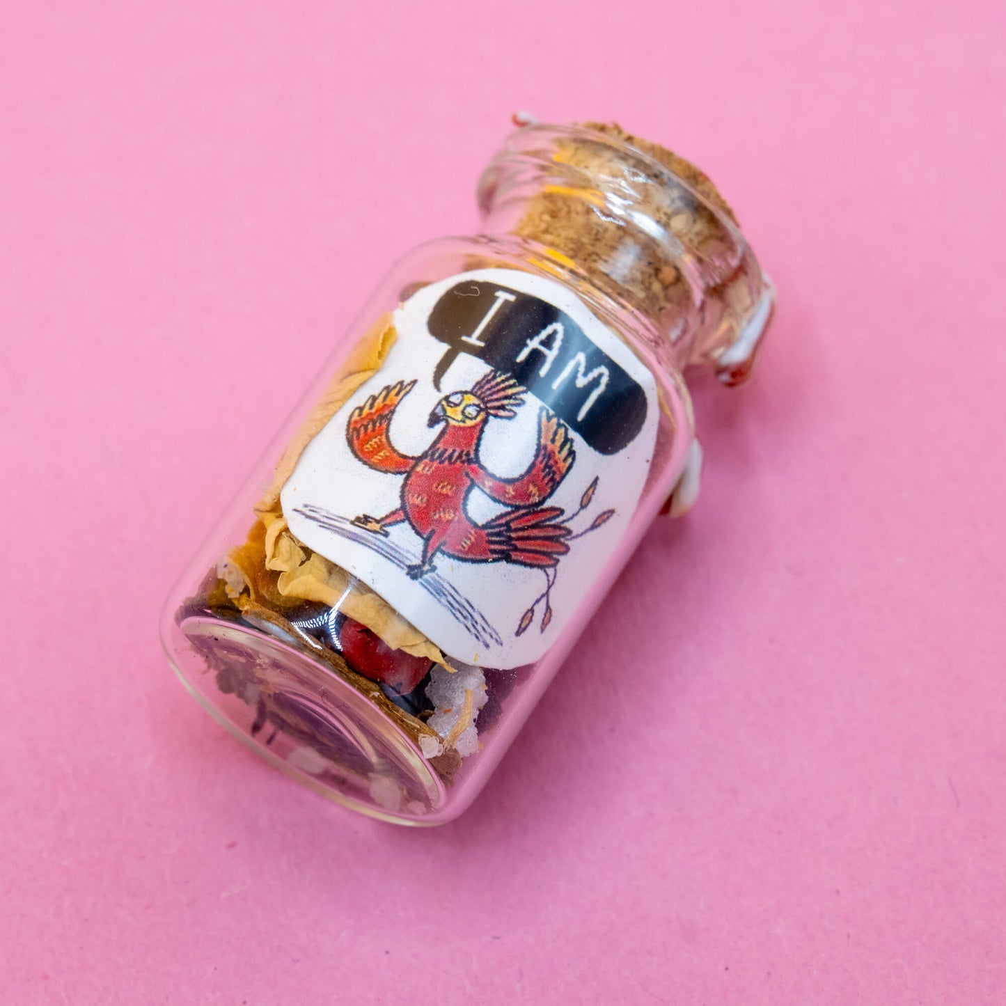 A handmade Phoenix-themed spell bottle lying on a pink surface, filled with magical herbs, petals, and crystals. The label shows a red phoenix character with a bold “I AM” affirmation. Sealed with cork and swirled wax. Created by Katie Abey & Angela Sandland as part of the Word Spells collection for confidence and self-empowerment.