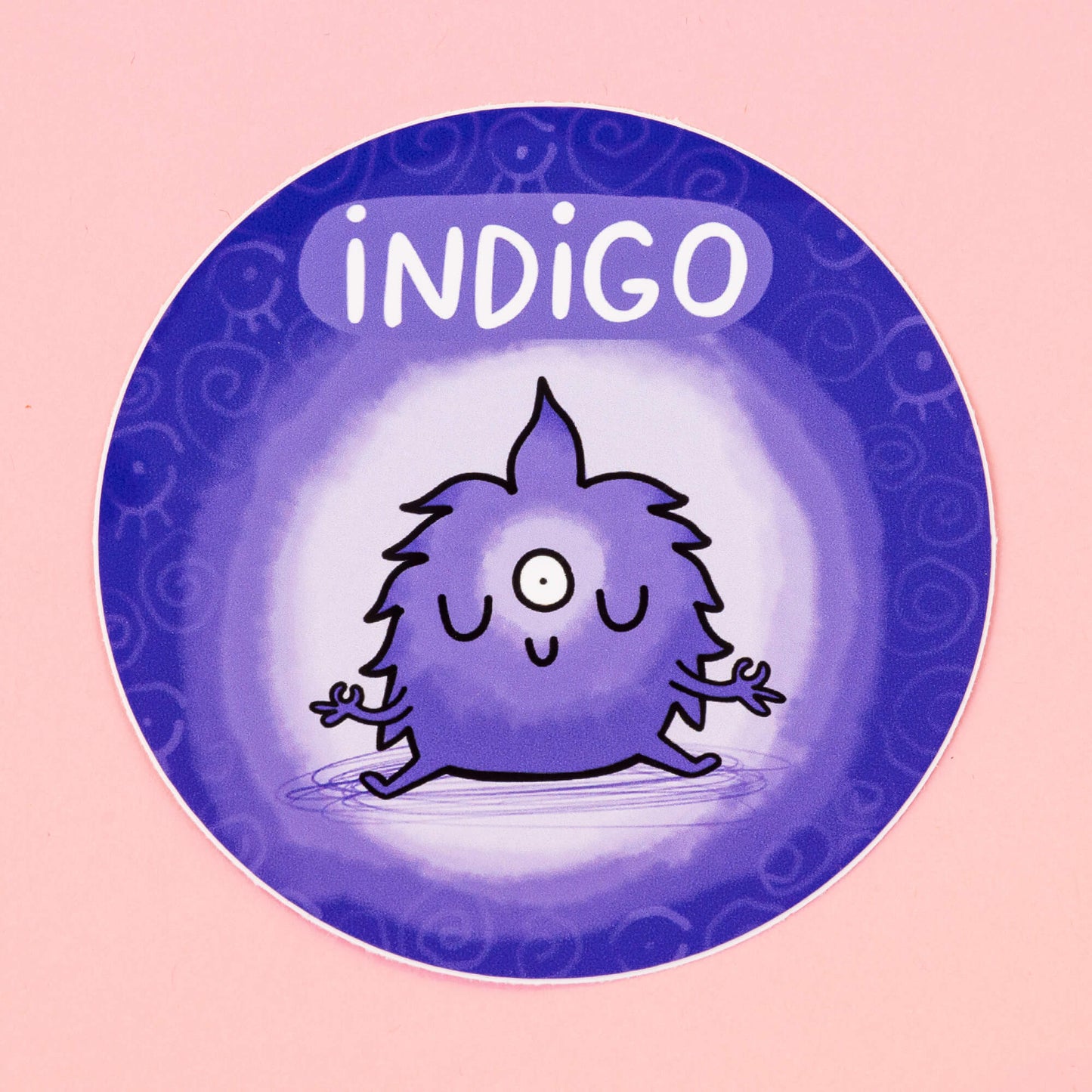 Indigo Chakreatures sticker by Happiness Enchanters. A circular indigo sticker with cute meditating cartoon creature with an open third eye illustrated by Katie Abey. 'Indigo' is written above the character. The sticker is on a pink backdrop.