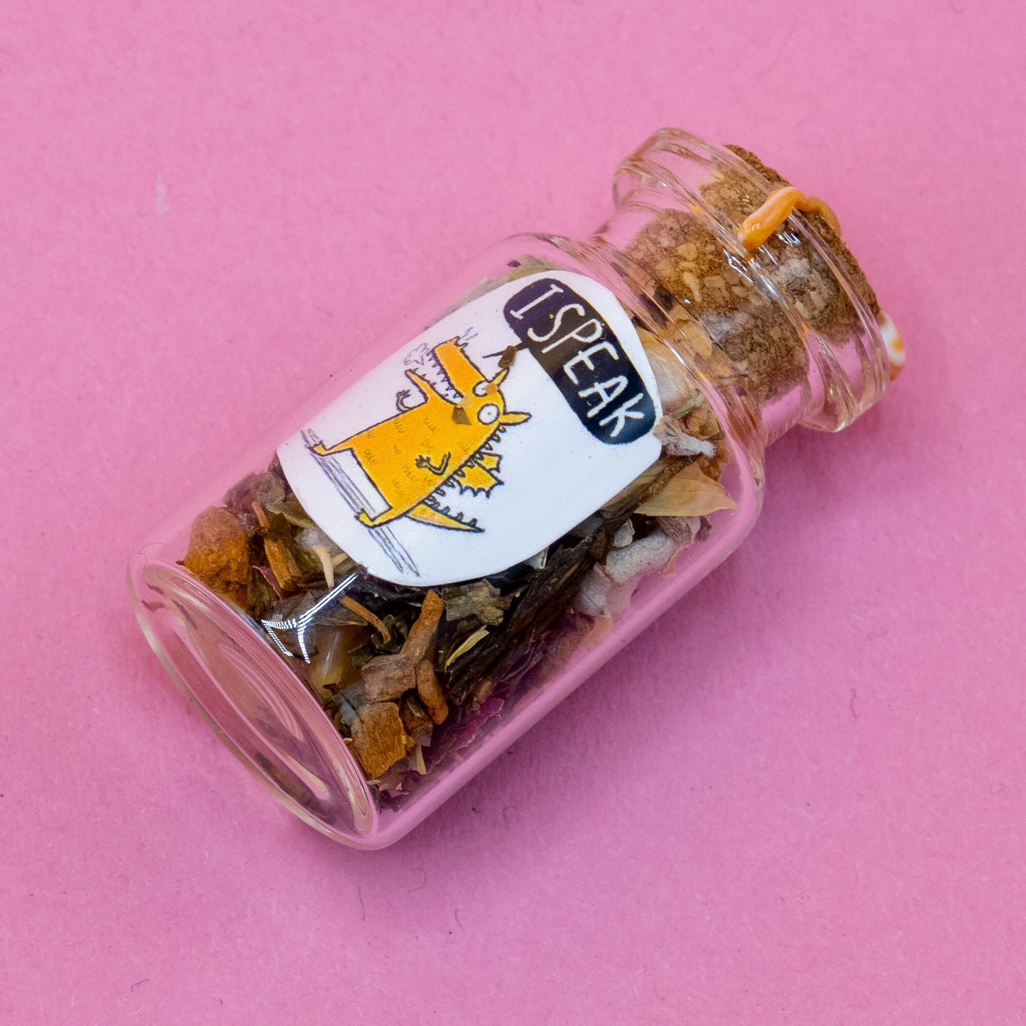 Glass spell bottle lying on a pink surface, filled with colourful herbs and botanical elements. Sealed with cork and yellow wax. The label features an orange dragon character with the affirmation “I SPEAK.” From the handmade Word Spells range by Katie Abey & Angela Sandland, designed to support clear communication and vocal confidence.
