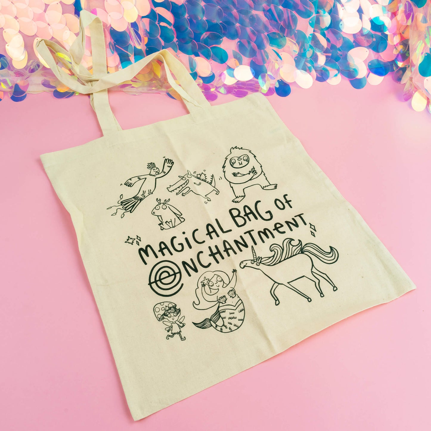 A whimsical cotton tote bag by Happiness Enchanters, titled "Magical Bag of Enchantment." The natural beige bag is illustrated with a collection of charming, hand-drawn magical creatures, including a unicorn, mermaid, mushroom fairy, friendly monsters, and a flying bird. The bag's playful and imaginative design offers a delightful, eco-friendly way to carry your essentials while adding a touch of magic to everyday errands. Set against a pastel pink background with shimmering holographic details