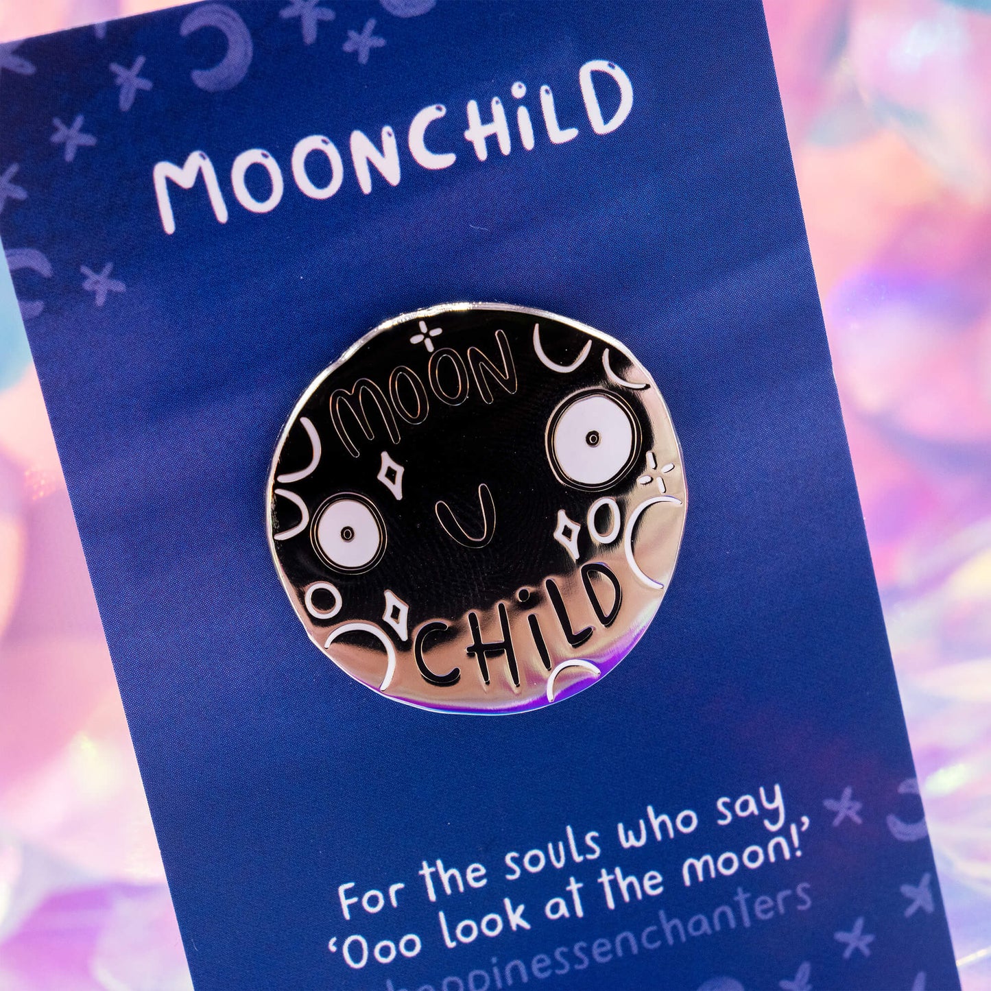 A close-up of the Moonchild pin badge by Happiness Enchanters, designed by Katie Abey. The silver shiny metal badge features a joyful moon illustration surrounded by stars and crescent shapes, glowing against a dreamy iridescent backdrop and blue backing card.