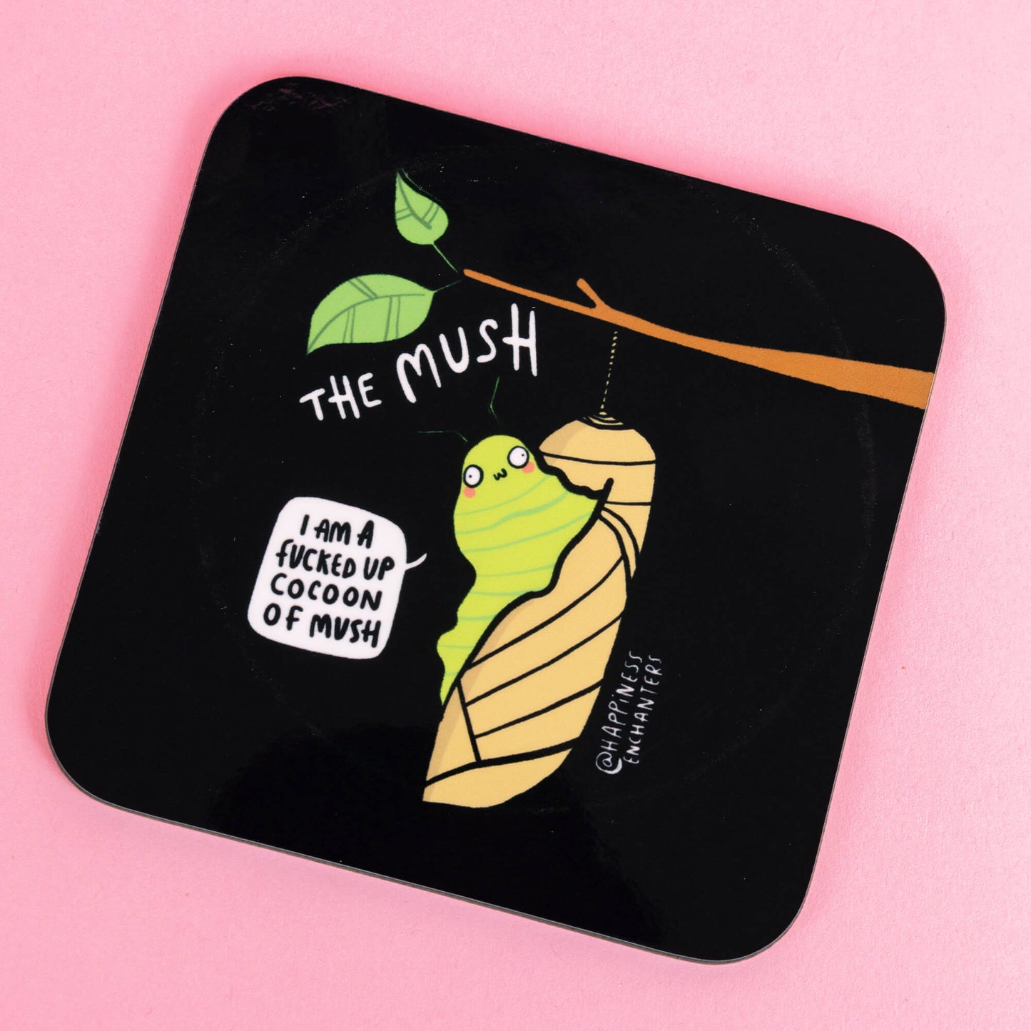 A glossy black coaster showing a quirky green caterpillar in a cocoon designed by Katie Abey. Text reads, "I am a f***ed up cocoon of mush," against a pastel pink background. A light-hearted creation by Happiness Enchanters, embracing transformation.