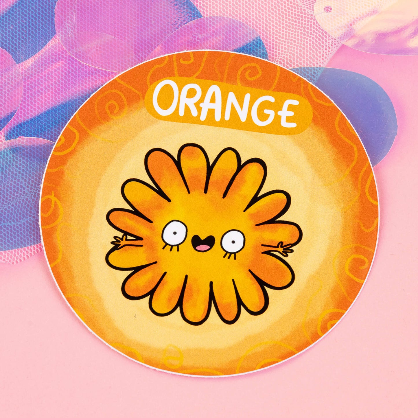Orange Chakreatures sticker by Happiness Enchanters. A circular sticker with an orange flower shaped creature illustrated by Katie Abey with outstretched arms, smiling mouth on a light orange background. 'Orange' is written above the character. The sticker is sat on a pink backdrop.