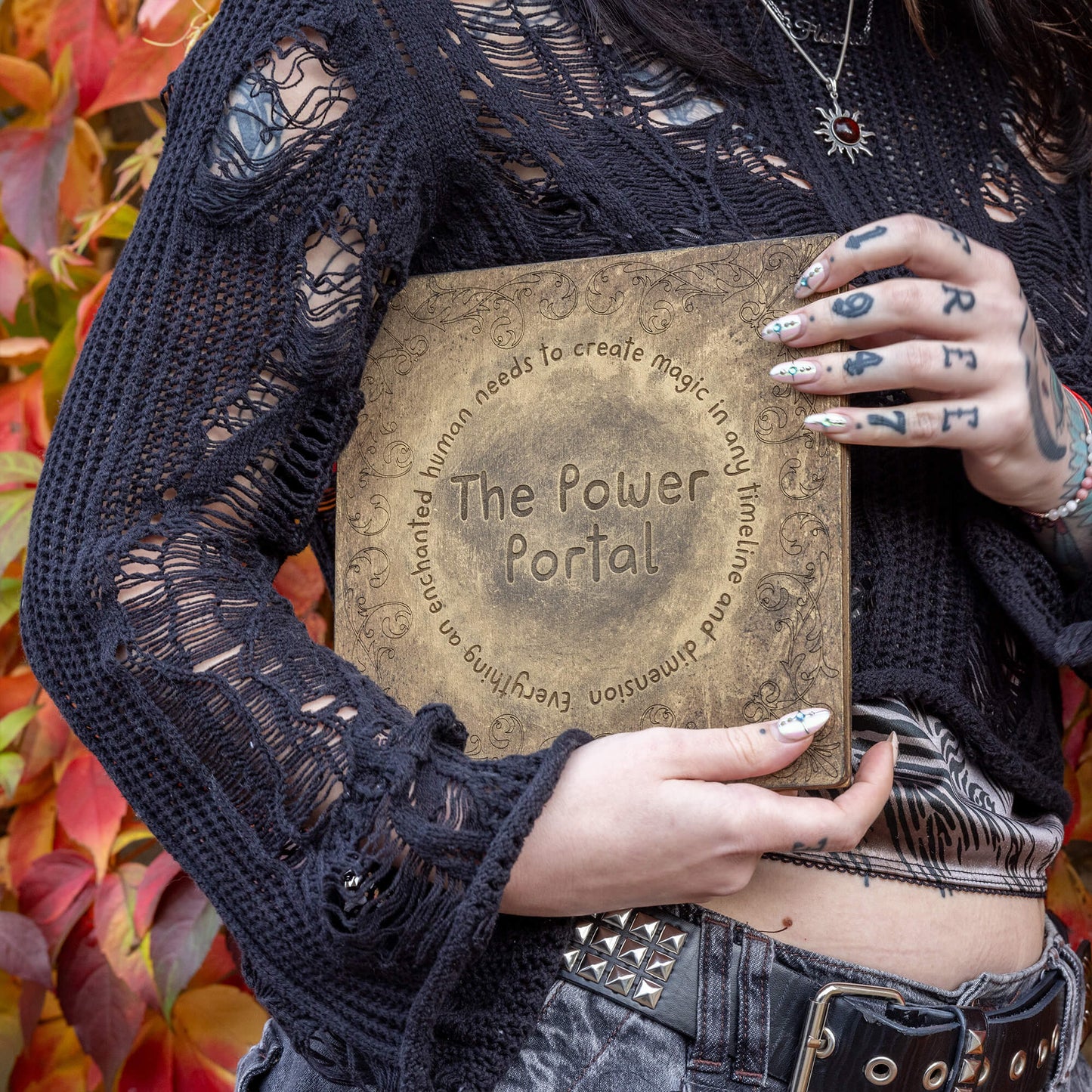 A person holding the Power Portal close to their chest, dressed in a black crochet top and decorated nails. The intricate engraving reads, "The Power Portal – everything an enchanted human needs to create magic in any timeline and dimension," capturing the magical essence of the Happiness Enchanters brand.