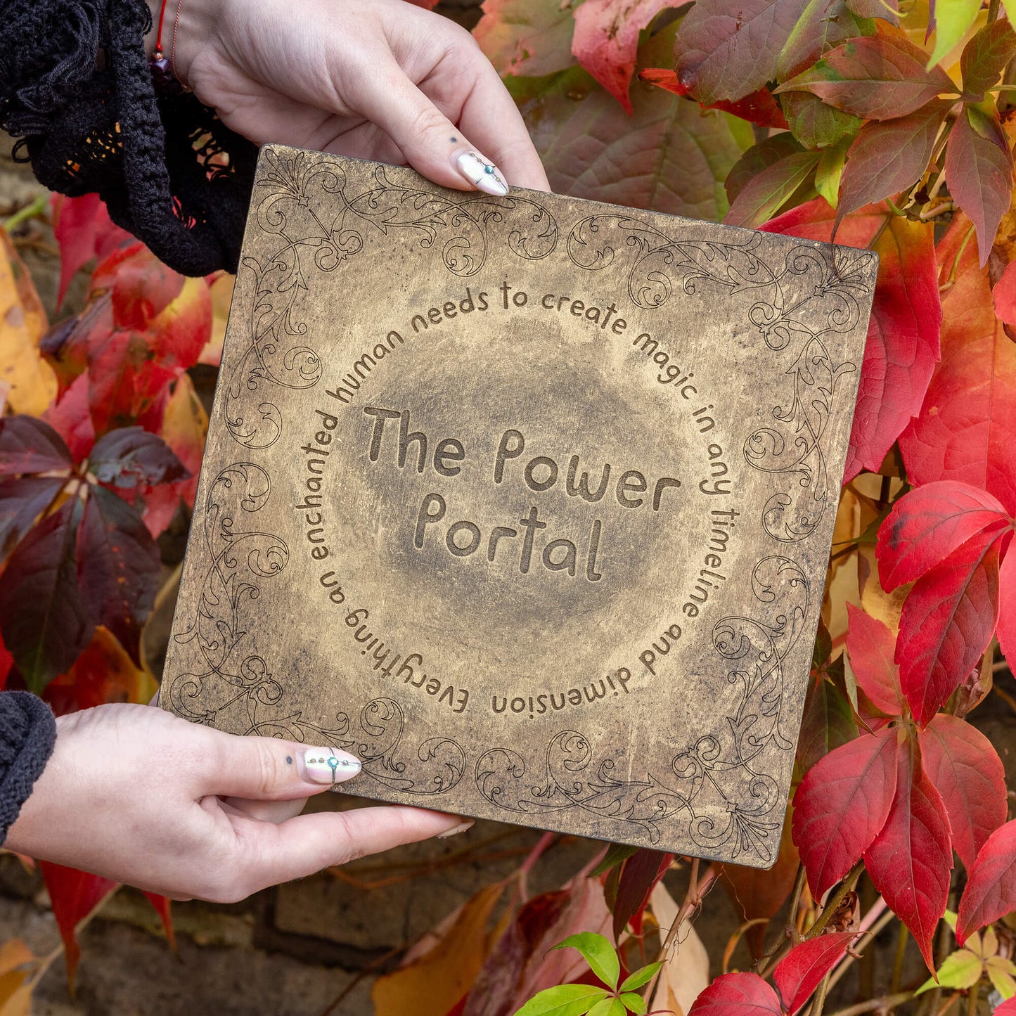 The Power Portal being gently held with two hands, surrounded by vibrant autumn leaves. The portal's bronze tone and engraved details reflect the natural, enchanted vibe of the brand. This mystical tool is used for connecting with magical energies