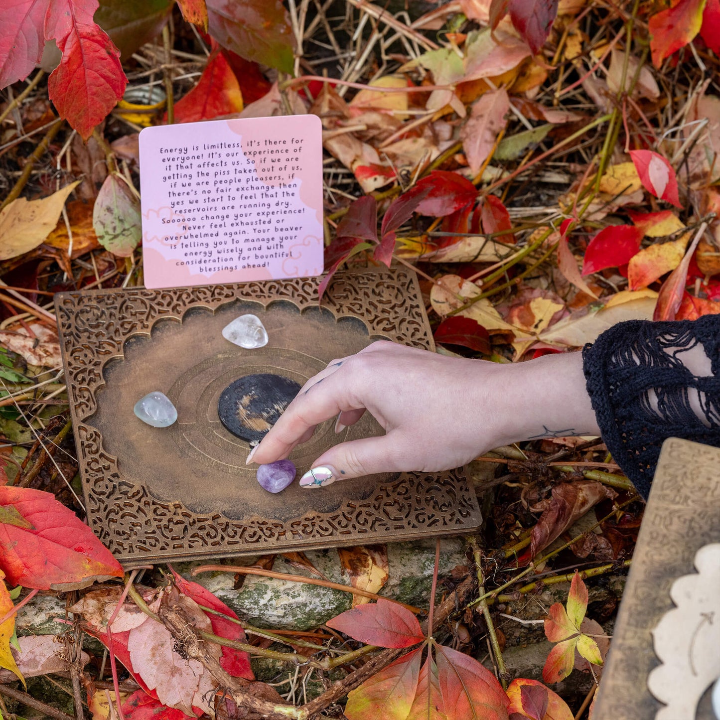 A hand placing an amethyst crystal on the Power Portal amidst autumn leaves and a card about managing energy. The delicate setup symbolises the practice of intention-setting and energy management in a magical, nature-infused space.