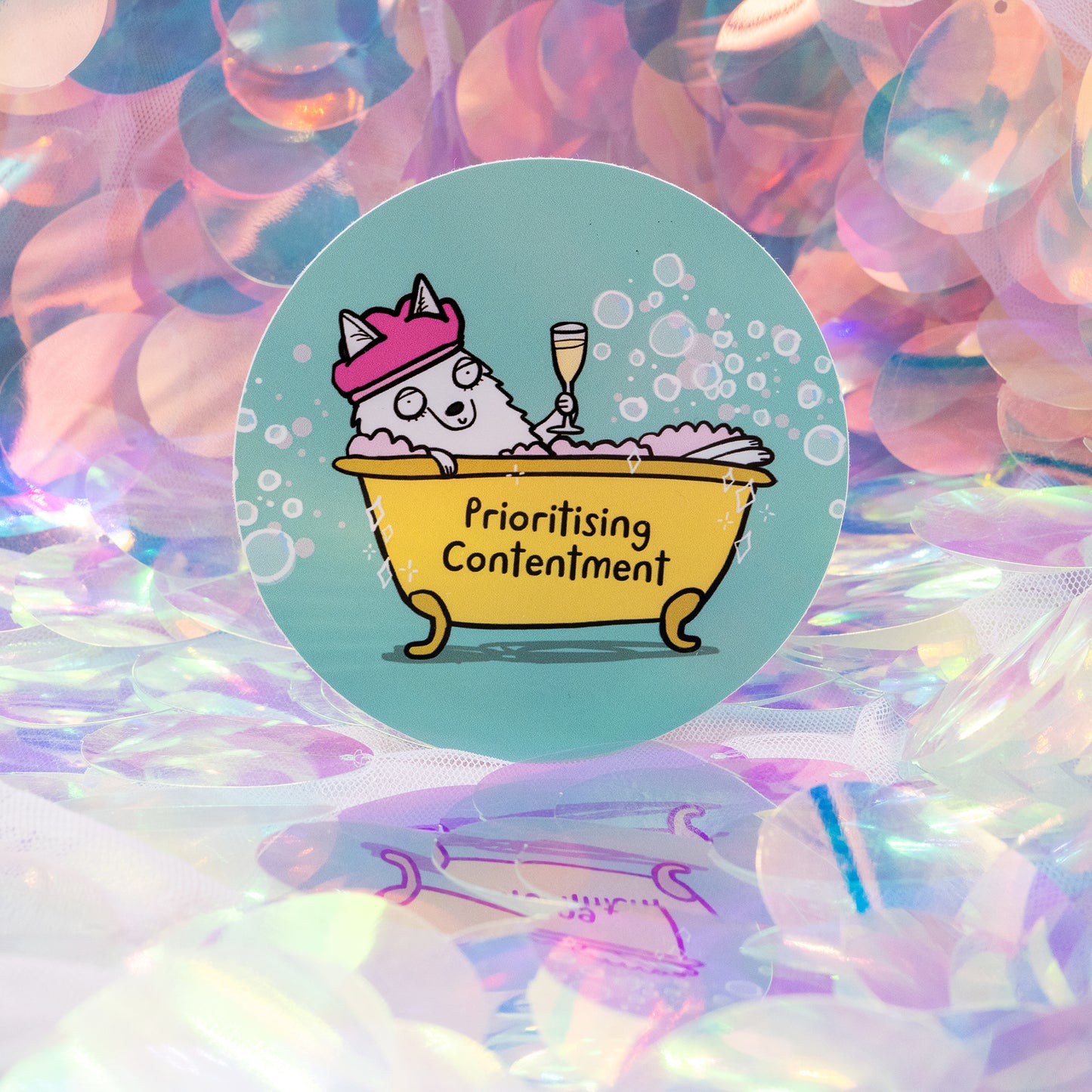 A round Happiness Enchanters vinyl sticker showing a cheerful illustrated wolf in a yellow bubble bath, wearing a pink shower cap and raising a champagne glass. The tub says “Prioritising Contentment”. Set against a sparkly iridescent backdrop. A whimsical Katie Abey design encouraging calm, joy and mindful self care.