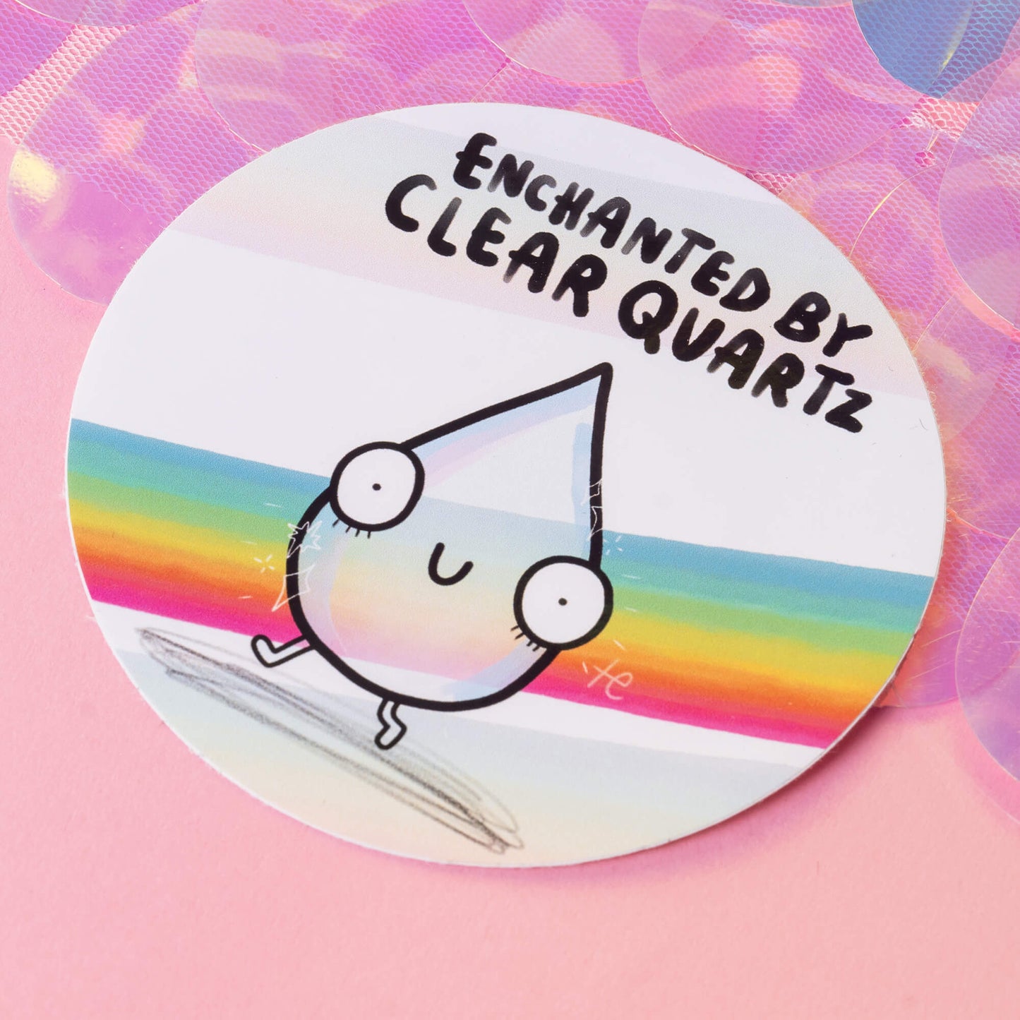 A round sticker with a whimsical, wide-eyed clear quartz crystal character on a colourful pastel rainbow background. The text above reads "Enchanted by Clear Quartz." The sticker is set against a pink background with iridescent circles, giving a magical and playful feel. Created by Happiness Enchanters, who inspire magic and creativity through art, illustration, writing, and more.