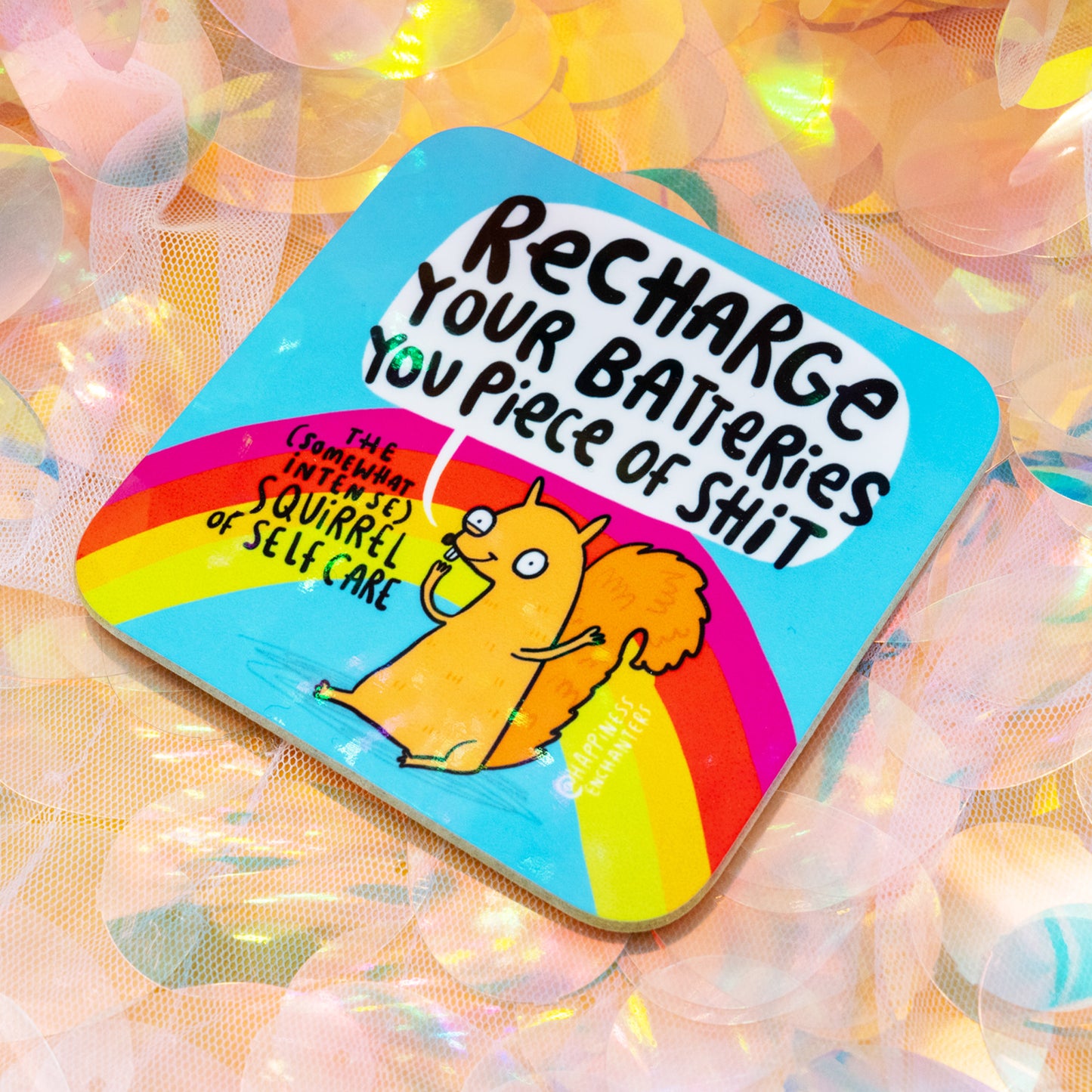A colourful square coaster featuring Katie Abey and Angela Sandland’s Happiness Enchanters design. It shows the “Somewhat Intense Squirrel of Self Care” holding a drink above a rainbow with the bold text “Recharge Your Batteries You Piece of Shit”. Shot on an iridescent sequin background. Fun self care gift coaster, bright rainbow design.