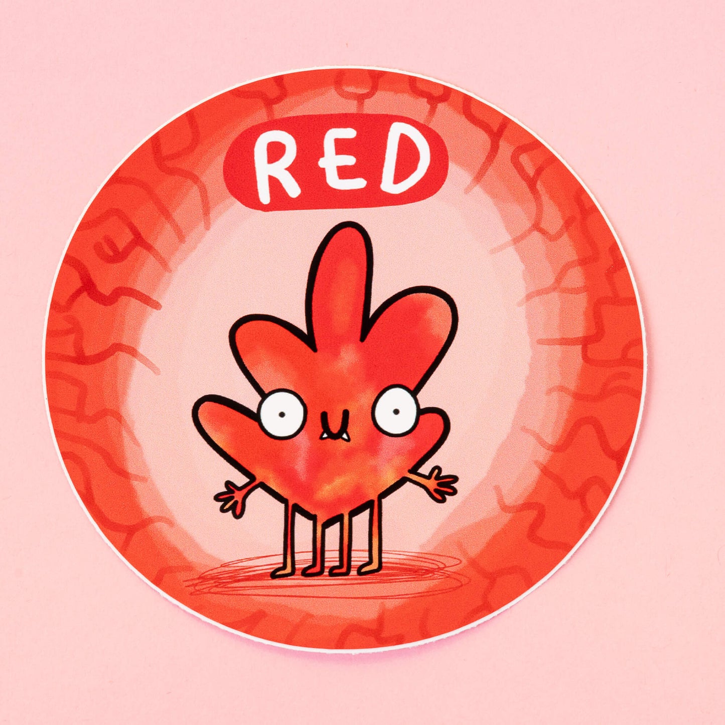 Red Chakreatures sticker by Happiness Enchanters. A circular sticker with a red leaf shaped creature illustrated by Katie Abey with outstretched arms, smiling mouth, four legs and fangs on a light orange background. 'Orange' is written above the character. The sticker is sat on a pink backdrop.