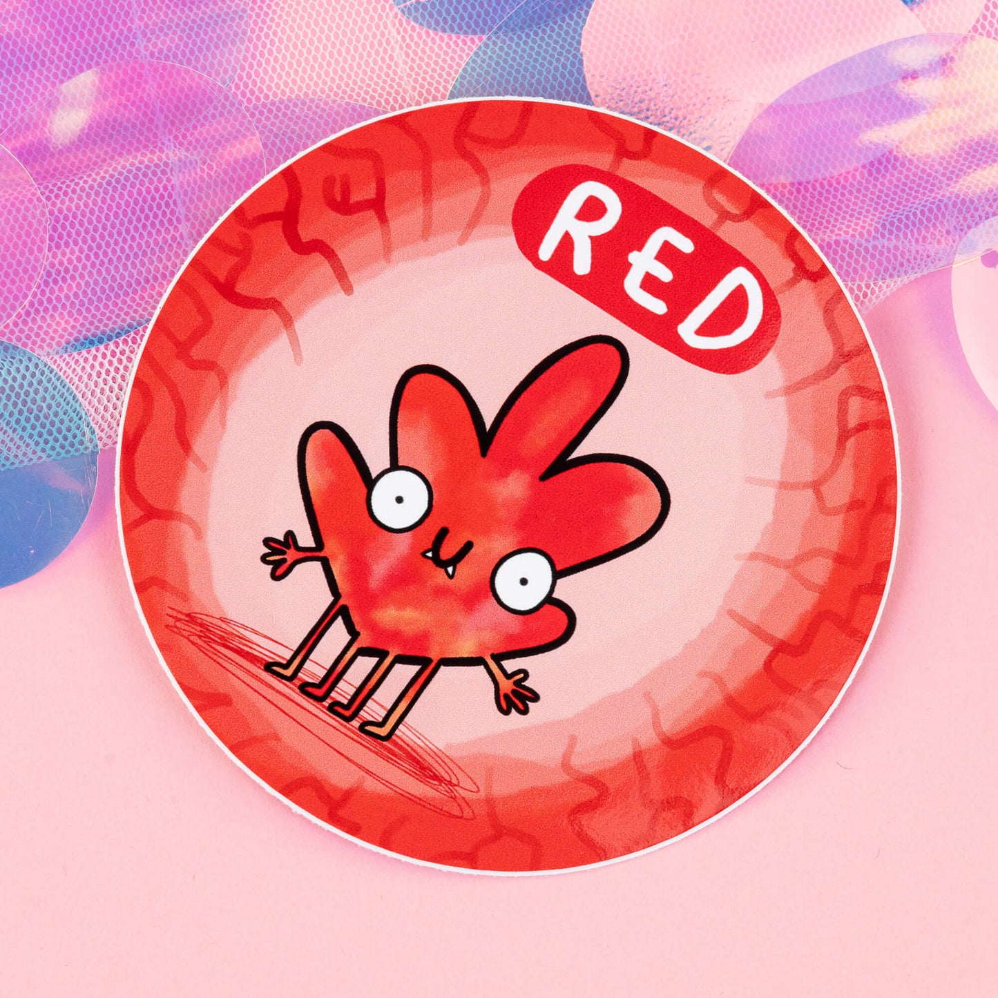 Red Chakreatures sticker by Happiness Enchanters. A circular sticker with a red leaf shaped creature illustrated by Katie Abey with outstretched arms, smiling mouth, four legs and fangs on a light orange background. 'Orange' is written above the character. The sticker is sat on a pink backdrop.