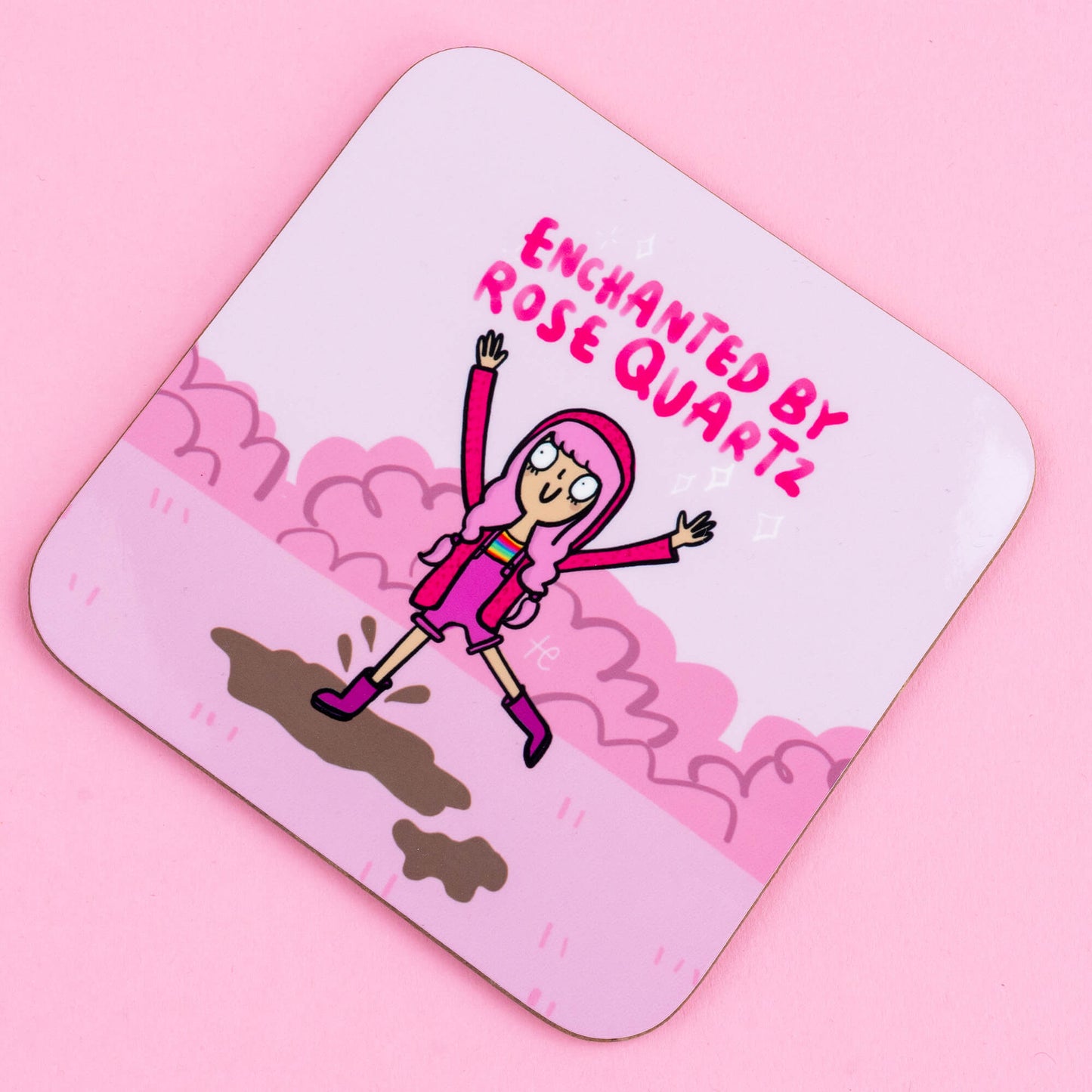 A close-up of a Happiness Enchanters coaster, showcasing a whimsical illustration of a pink-haired character jumping with arms raised, surrounded by pink clouds. The text "Enchanted by Rose Quartz" adds a playful and enchanting vibe, reflecting the brand's creative and mystical aesthetic.