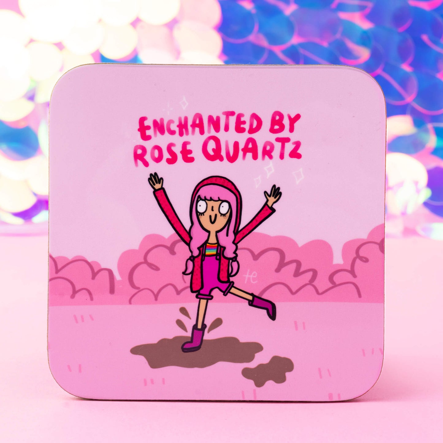 A vibrant coaster design from Happiness Enchanters, featuring a cheerful cartoon character with pink hair, a matching pink outfit, and boots, joyfully leaping in a pink landscape under the words "Enchanted by Rose Quartz." The background is filled with pink clouds, enhancing the magical theme.