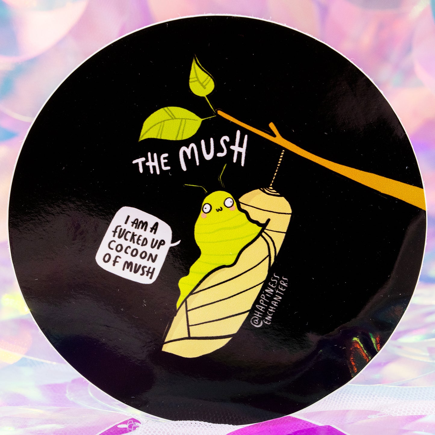 Close-up of The Mush Sticker by Happiness Enchanters. The design shows a cute green caterpillar halfway in a cocoon on a branch, saying “I am a fucked up cocoon of mush”, with “The Mush” written above. Displayed on a glossy black circle with an iridescent pastel backdrop for a whimsical, relatable feel.