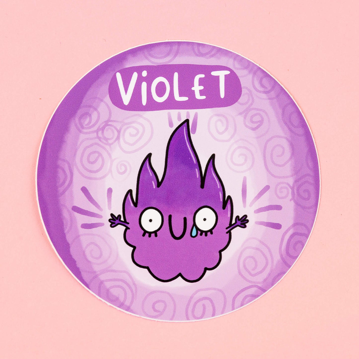 Violet Chakreatures sticker by Happiness Enchanters. A circular sticker with a violet flame shaped creature illustrated by Katie Abey with outstretched arms, smiling mouth a tear in it's eye on a light purple background. 'Violet' is written above the character. The sticker is sat on a pink backdrop.
