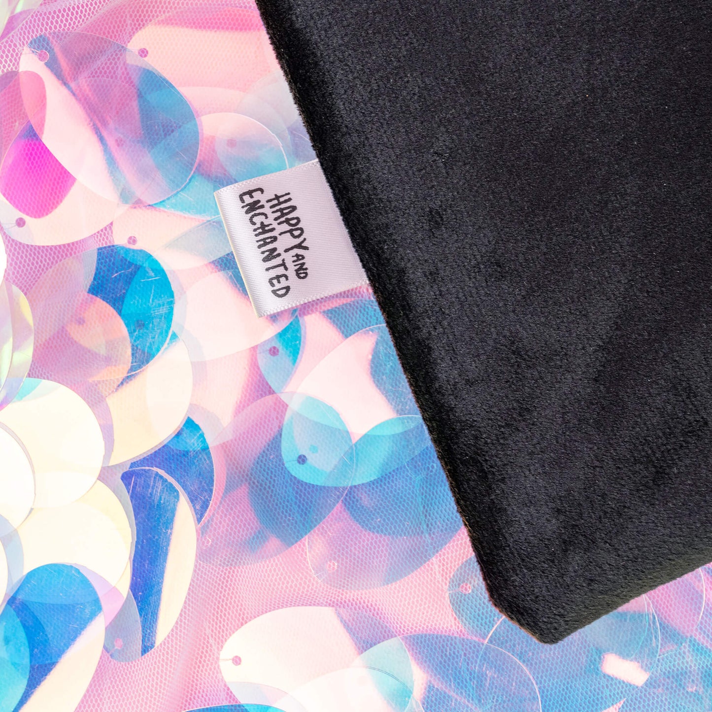 Detail of the Happiness Enchanters black velvet bag featuring a rainbow stripe and a branded tag reading ‘HAPPY AND ENCHANTED.’ The bag lies on a background of iridescent sequins, reflecting the brand’s commitment to magic and creativity.