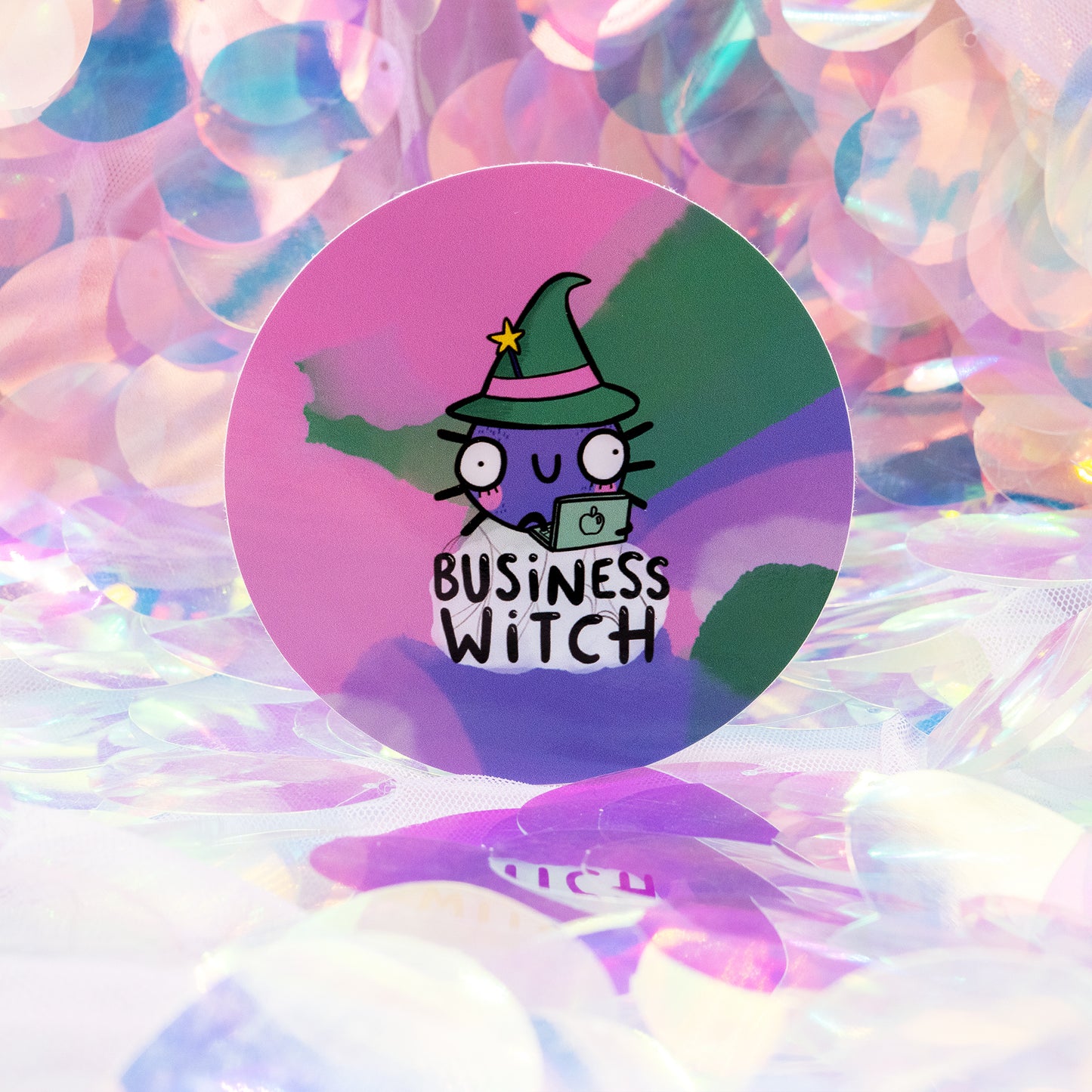 A round “Business Witch” sticker by Happiness Enchanters, created by Katie Abey and Angela Sandland. The sticker features a cute purple witch character in a green hat working on a laptop, set against a pastel pink and rainbow background. Photographed on an iridescent, shimmery surface for a magical, whimsical look.