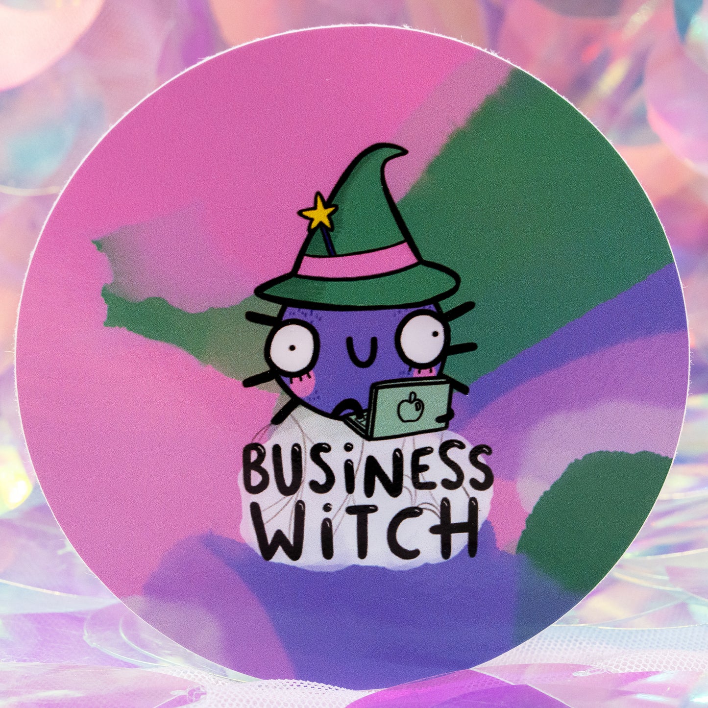 Close up of the round “Business Witch” sticker by Happiness Enchanters, illustrated by Katie Abey and Angela Sandland. Shows a purple witch character in a green hat using a laptop, with bold “Business Witch” text underneath. Set on a colourful pastel gradient background with soft, dreamy lighting for a magical feel.