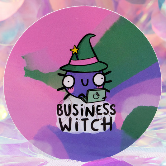 Close up of the round “Business Witch” sticker by Happiness Enchanters, illustrated by Katie Abey and Angela Sandland. Shows a purple witch character in a green hat using a laptop, with bold “Business Witch” text underneath. Set on a colourful pastel gradient background with soft, dreamy lighting for a magical feel.