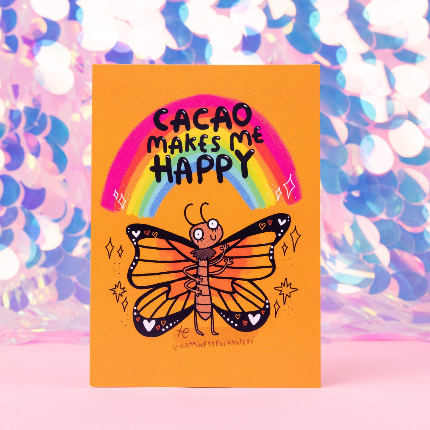 An orange postcard with a butterfly illustration by Katie Abey with sparkles and a rainbow, text reads Cacao makes me happy