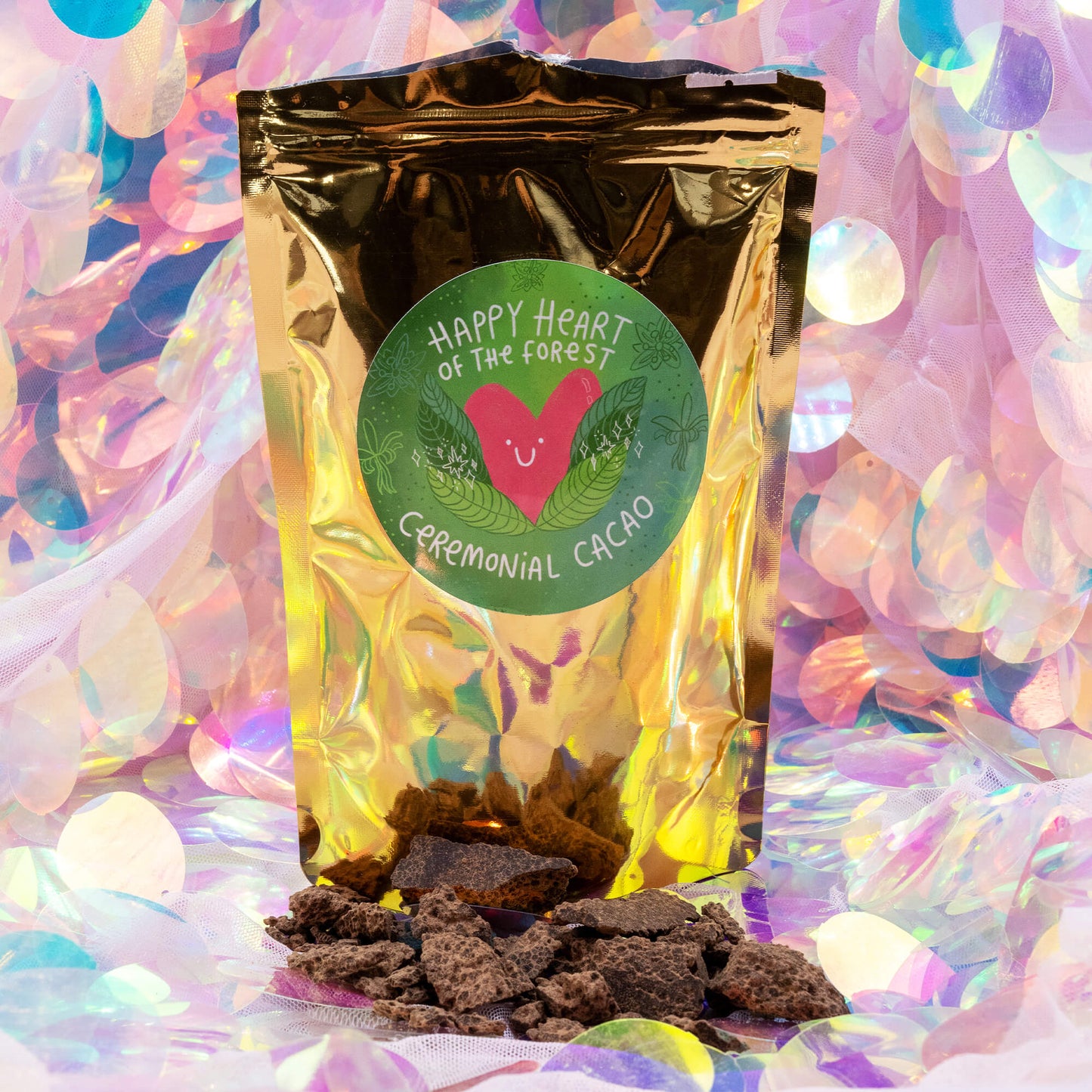Gold pouch of Happy Heart of the Forest Ceremonial Cacao by Happiness Enchanters surrounded by scattered cacao pieces. The packaging features a smiling heart design with green leaves.