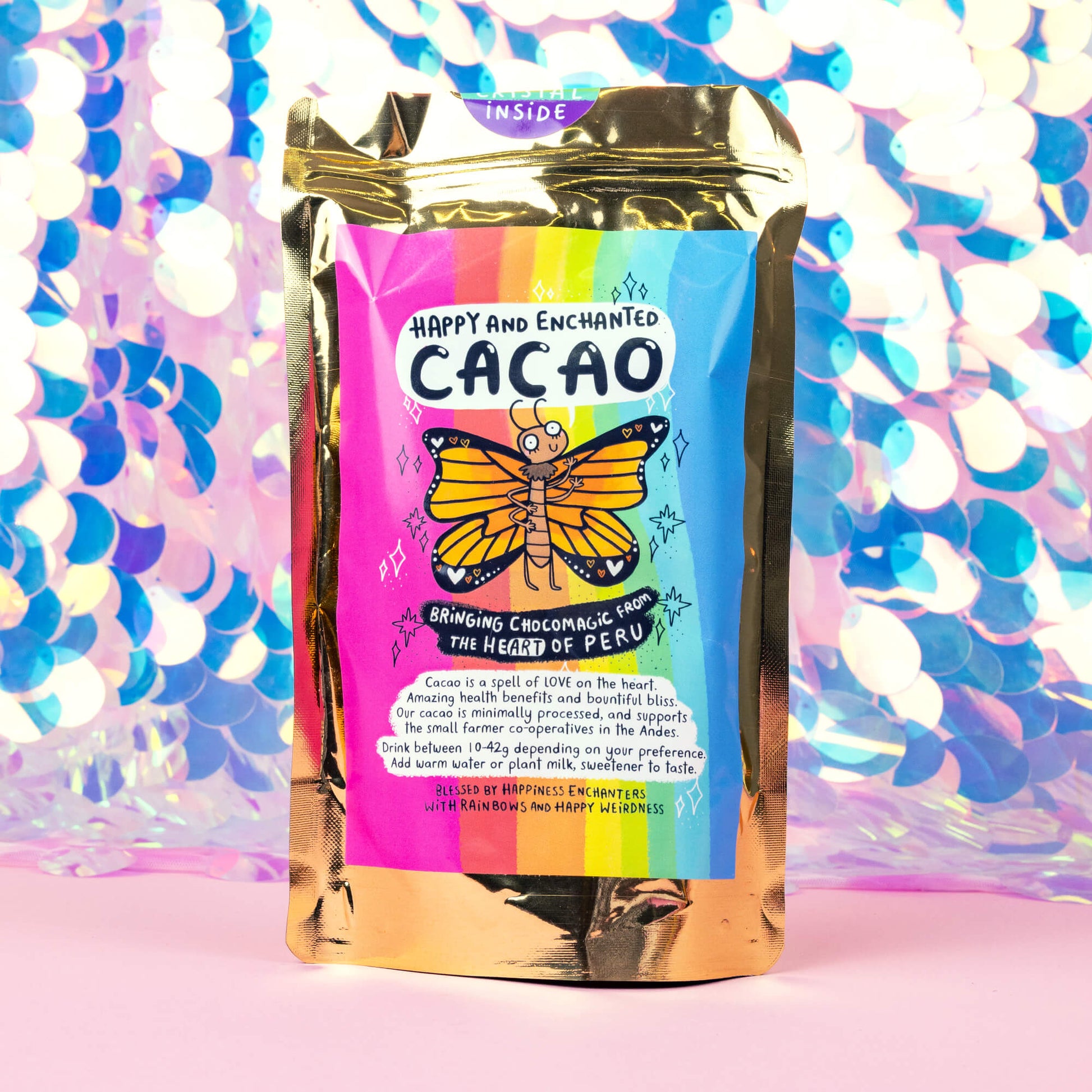 A colorful package of “Happy and Enchanted Cacao” with a rainbow design and an illustration of a butterfly is displayed against a vibrant, shiny background. The package is gold with a label indicating it brings “ChocoMagic from the heart of Peru.” In front of the package, there are cacao nibs and a blue crystal on a pink surface.