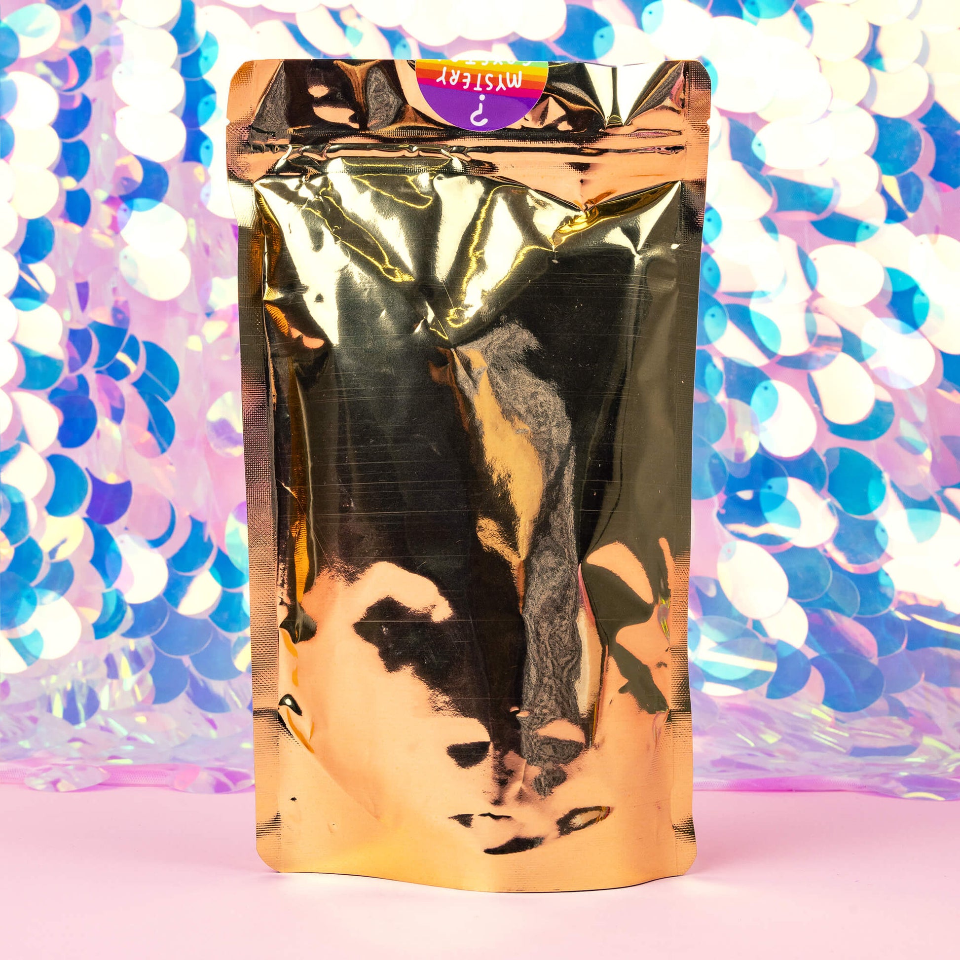 A colorful package of “Happy and Enchanted Cacao” with a rainbow design and an illustration of a butterfly is displayed against a vibrant, shiny background. The package is gold In front of the package