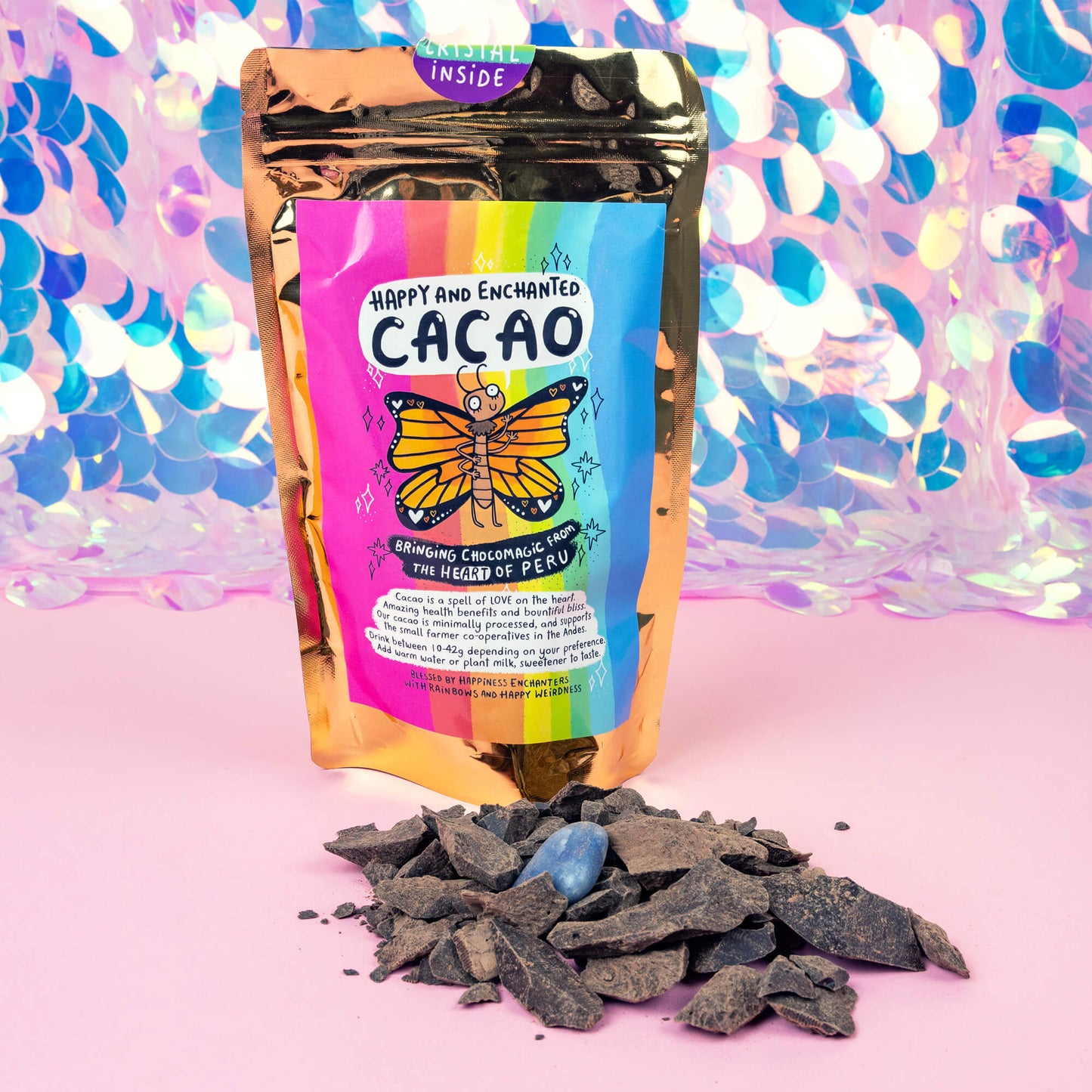 A colorful package of “Happy and Enchanted Cacao” with a rainbow design and an illustration of a butterfly is displayed against a vibrant, shiny background. The package is gold with a label indicating it brings “ChocoMagic from the heart of Peru.” In front of the package, there are cacao nibs and a blue crystal on a pink surface.