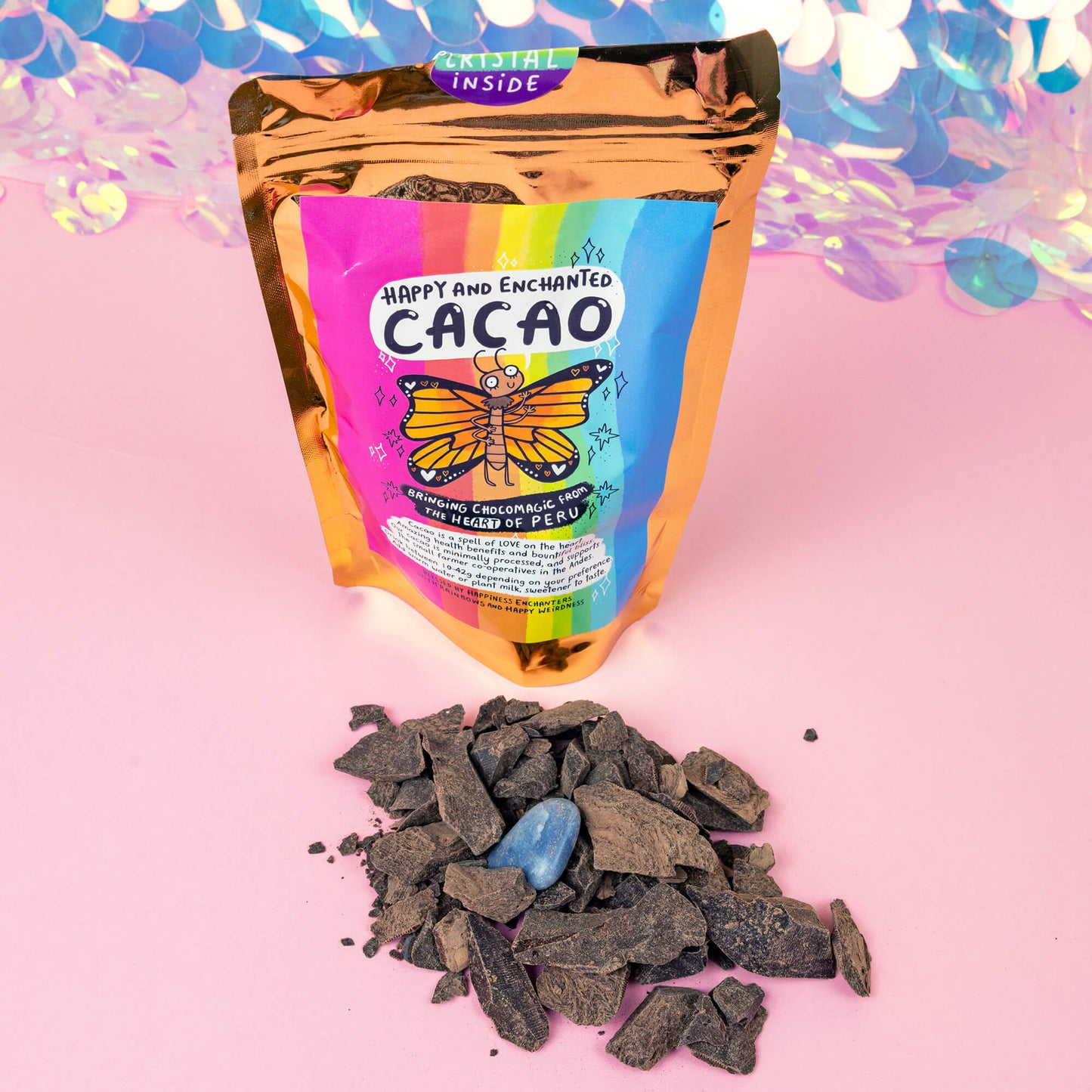 A colorful package of “Happy and Enchanted Cacao” with a rainbow design and an illustration of a butterfly is displayed against a vibrant, shiny background. The package is gold with a label indicating it brings “ChocoMagic from the heart of Peru.” In front of the package, there are cacao nibs and a blue crystal on a pink surface.