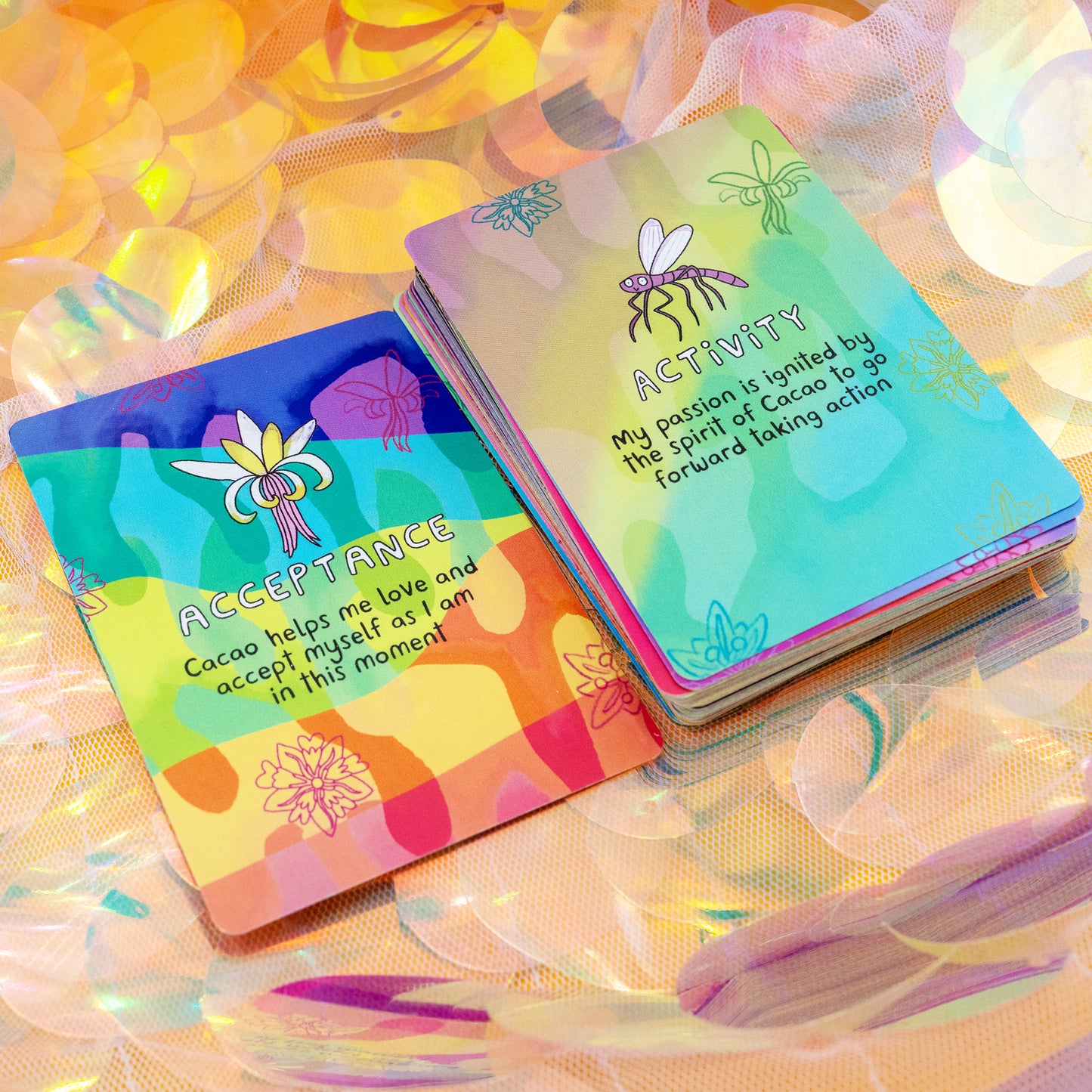 Two Cacao Journey Cards by Happiness Enchanters on iridescent fabric, showing the “Acceptance” and “Activity” cards with colourful illustrations. Created by Katie Abey and Angela Sandland to support cacao ceremonies, meditation and mindful daily practice.