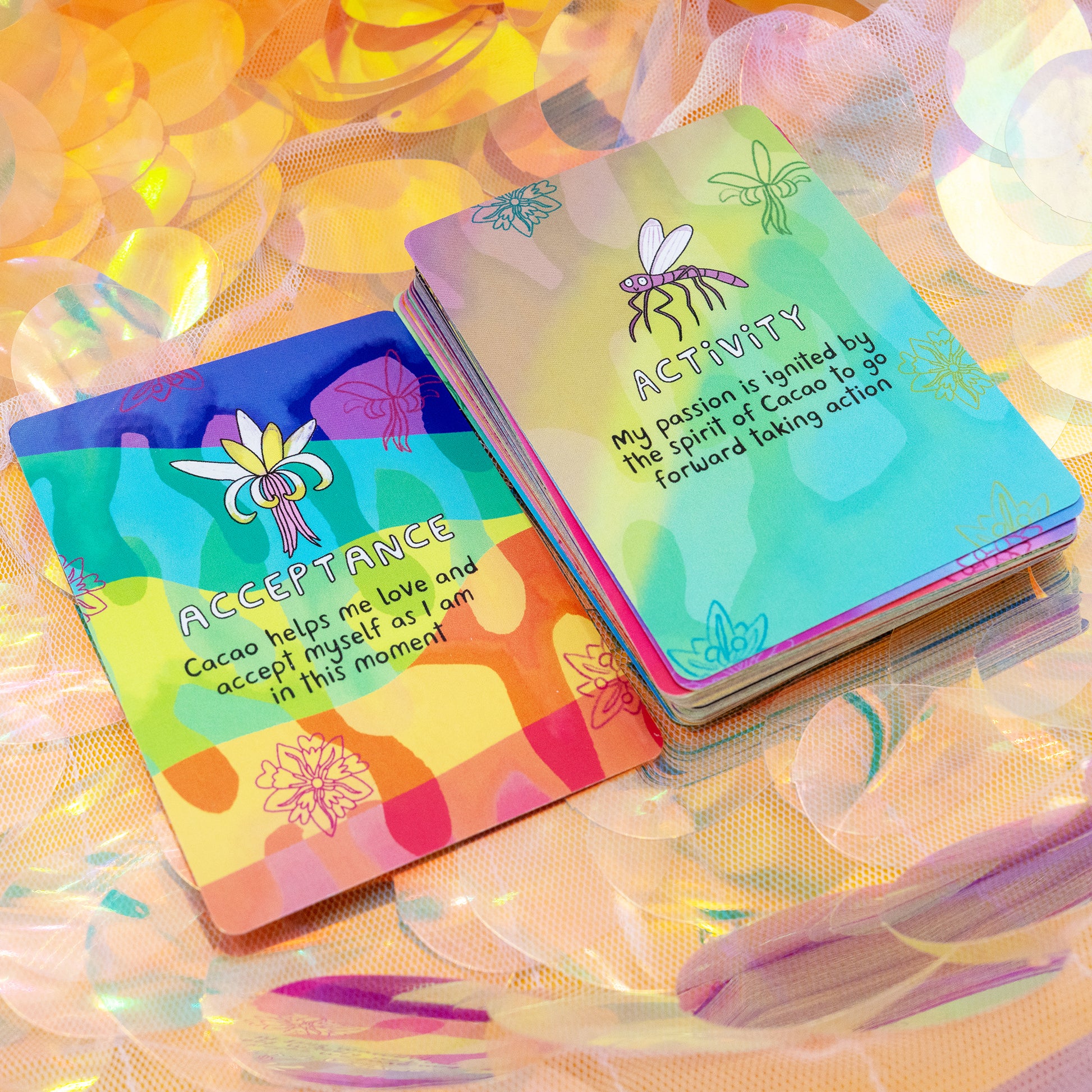Two Cacao Journey Cards by Happiness Enchanters on iridescent fabric, showing the “Acceptance” and “Activity” cards with colourful illustrations. Created by Katie Abey and Angela Sandland to support cacao ceremonies, meditation and mindful daily practice.