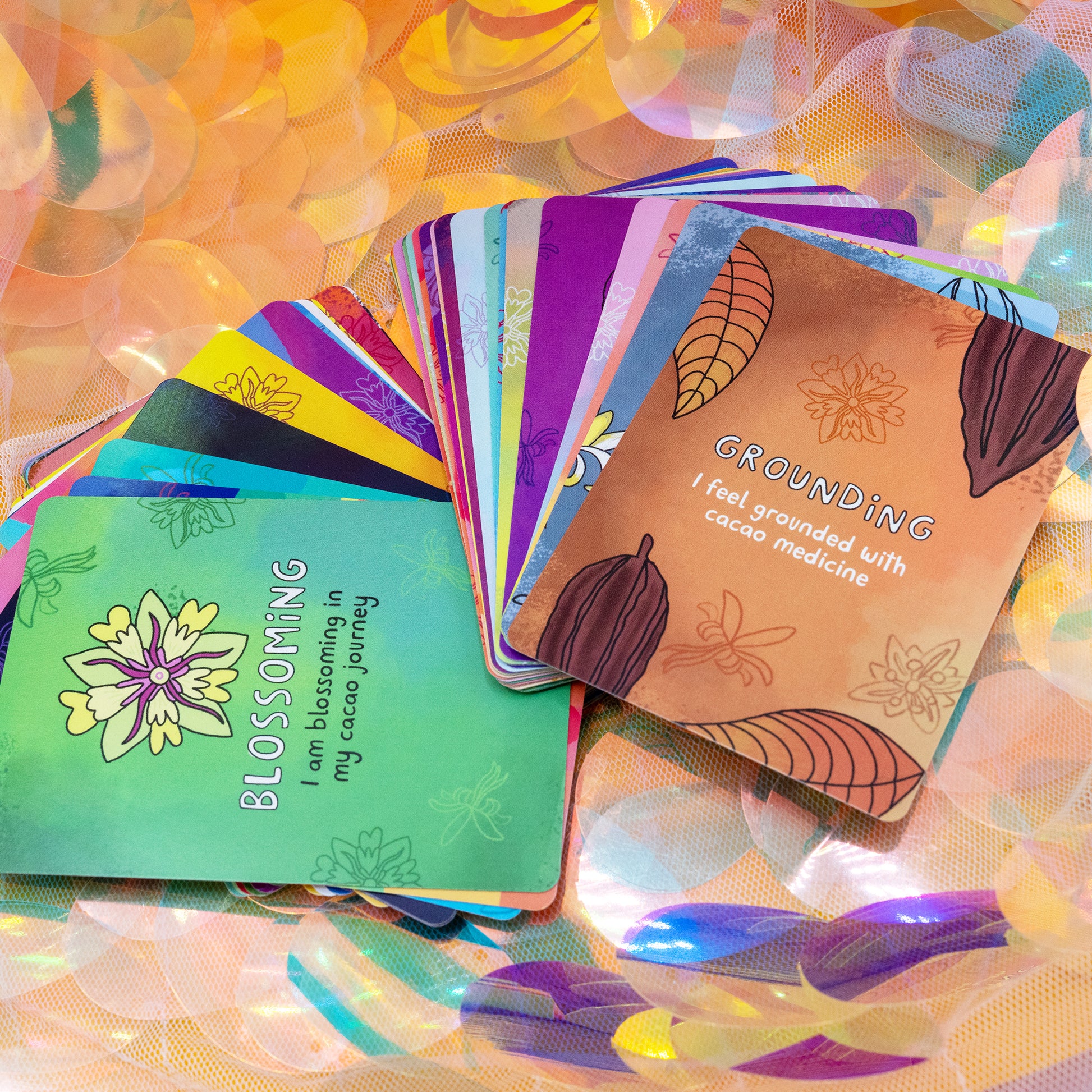 A colourful fan of Cacao Journey Cards by Happiness Enchanters with the “Grounding” card on top, featuring cacao pod illustrations. Designed by Katie Abey and Angela Sandland, these vibrant affirmation cards are used for cacao ceremonies, meditation and plant medicine guidance.