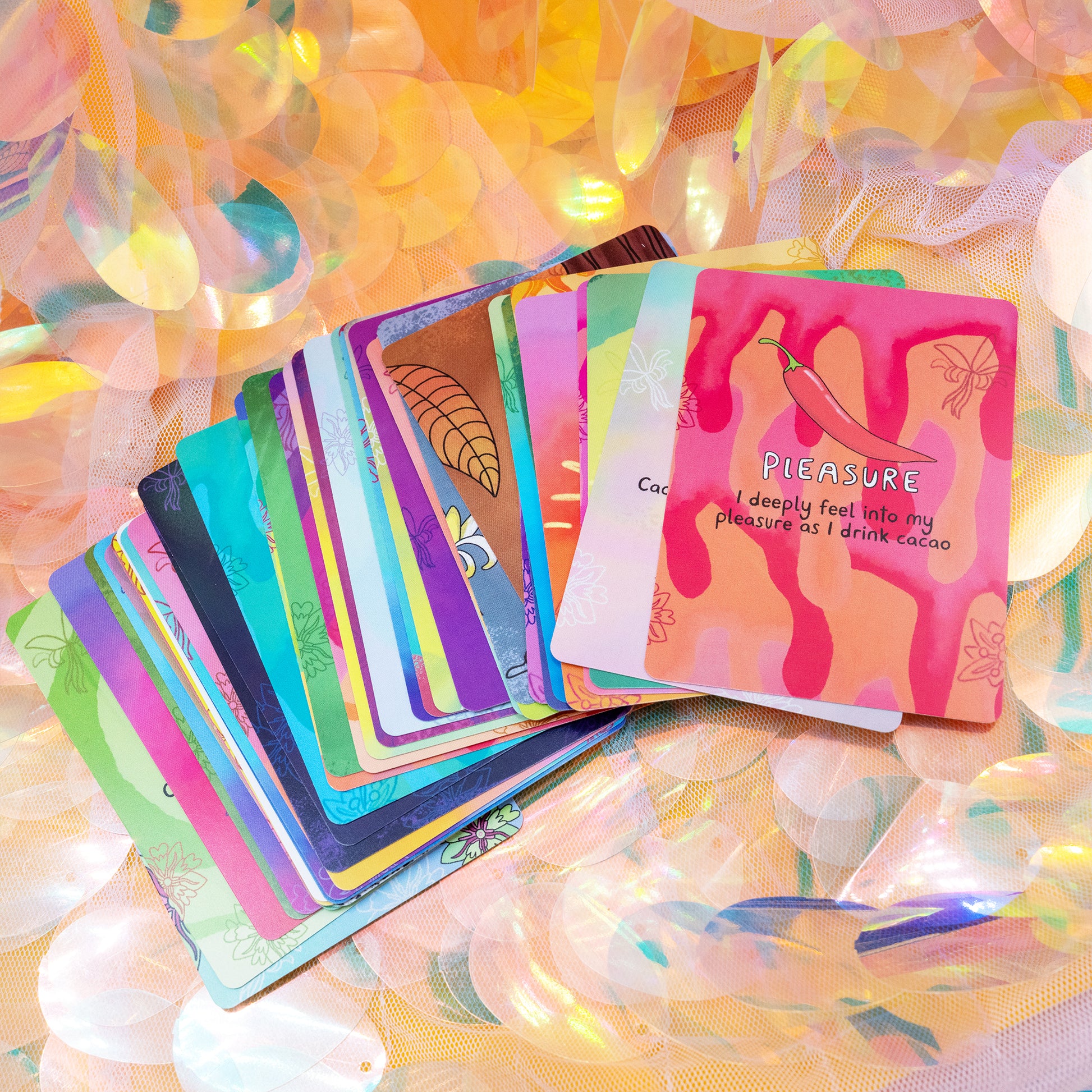 A spread of Cacao Journey Cards by Happiness Enchanters on iridescent fabric, showing bright illustrated designs and an affirmation card reading “Pleasure – I deeply feel into my pleasure as I drink cacao”. Created by Katie Abey and Angela Sandland for cacao rituals, mindfulness and spiritual reflection.