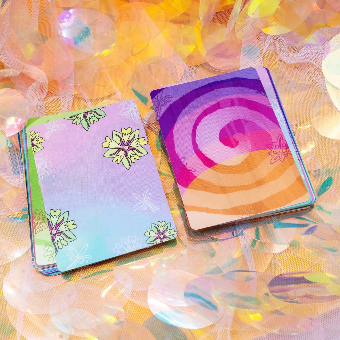 A colourful set of Cacao Journey Cards by Happiness Enchanters displayed on shimmering iridescent fabric. The cards feature vibrant illustrated backs with flowers and spirals. Created by Katie Abey and Angela Sandland, these oracle style cards support cacao rituals, meditation and mindful plant medicine journeys.