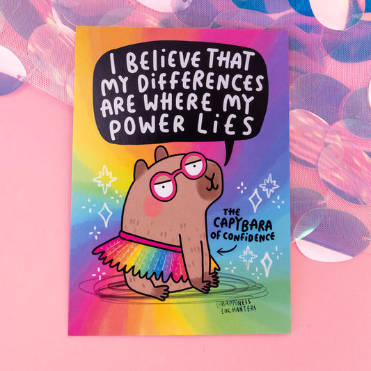 A whimsical postcard from 'Happiness Enchanters' featuring a cartoon capybara wearing pink glasses and a rainbow tutu, with the text 'I believe that my differences are where my power lies' on a vibrant rainbow background. The postcard exudes a magical and playful vibe, with the caption 'The Capybara of Confidence'.