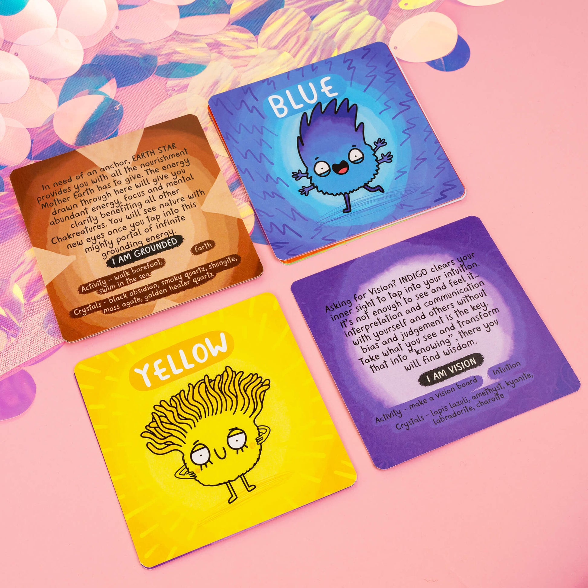 Four Chakreatures cards from Happiness Enchanters, each featuring a colourful character representing a different chakra. Cards include descriptions and affirmations like "I am Grounded" and "I am Vision," along with crystal and activity recommendations for chakra alignment. Set against a pink background with iridescent accents