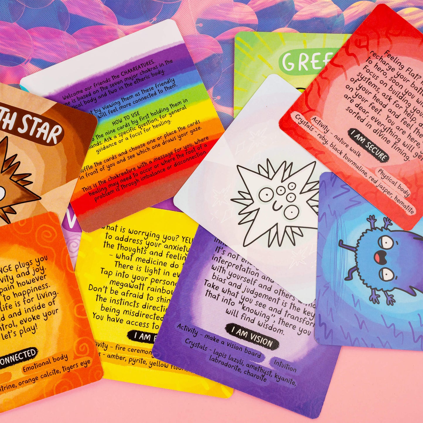 A spread of seven Chakreatures cards, each representing a chakra with a vibrant character, along with a rainbow instruction card explaining how to use the deck. Each card provides guidance, crystals, and activities for balancing chakras. Displayed on a magical pink and purple background