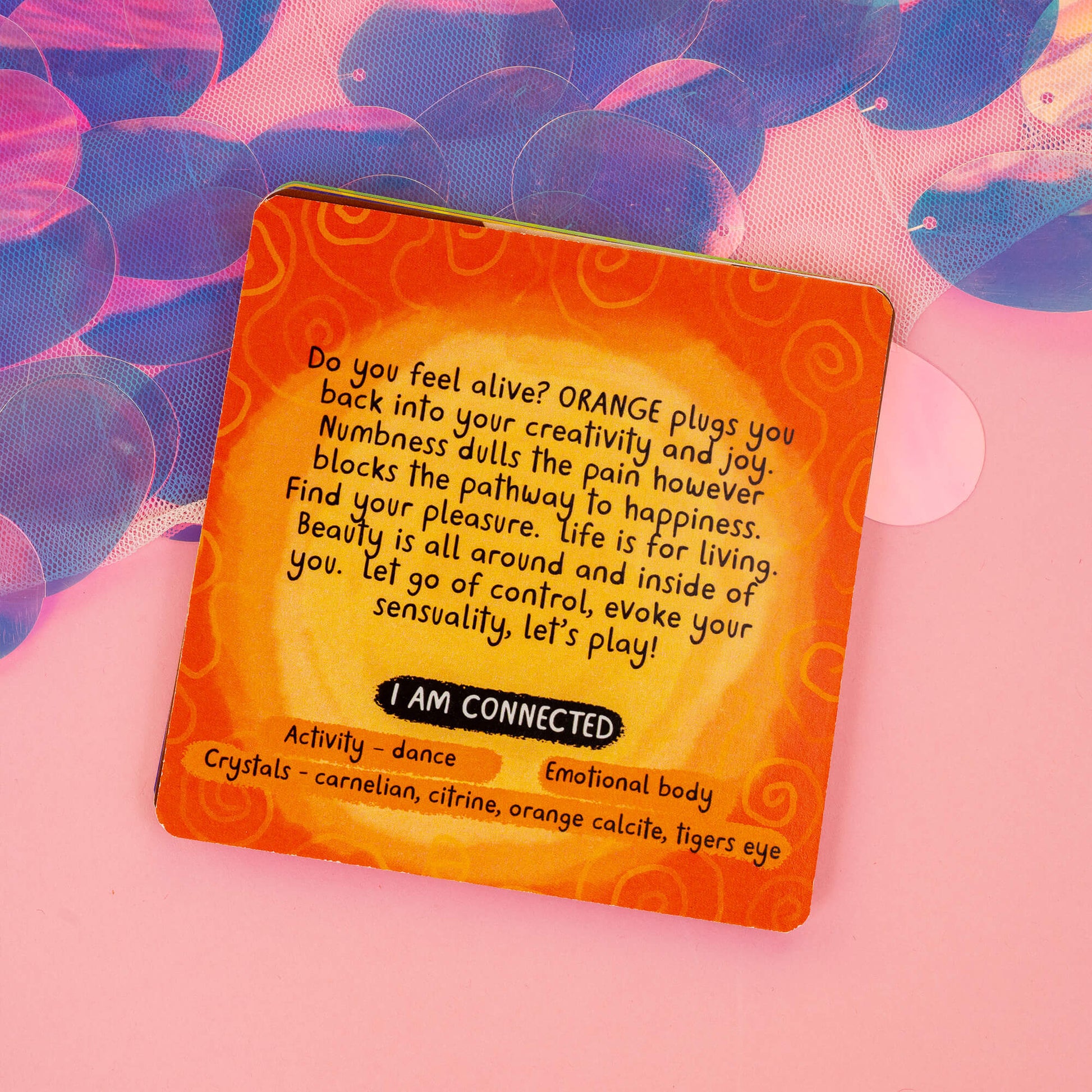 Close-up of the "Orange" chakra card from the Chakreatures deck by Happiness Enchanters. The card’s description encourages creativity, joy, and sensuality, with activities like dance and recommended crystals such as carnelian and citrine. Set on a pink background with holographic elements